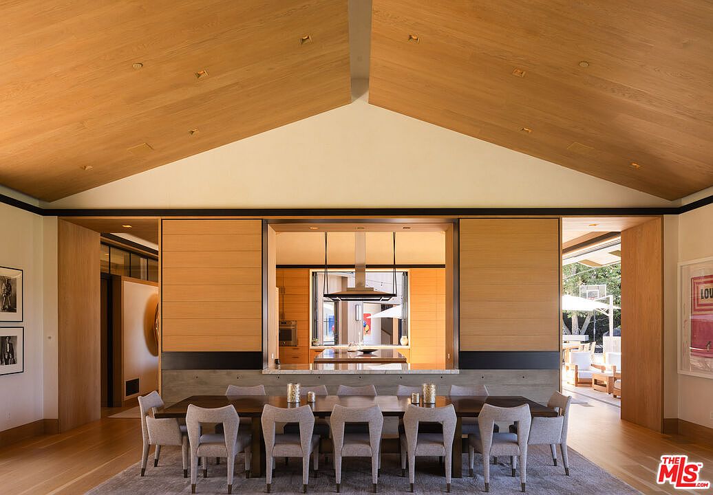 This elegant dining room features a high, vaulted wood-paneled ceiling that creates a grand, airy atmosphere. A long, dark wood table is surrounded by eight upholstered chairs, positioned in front of a large opening that looks through to a modern kitchen and an outdoor patio area. The space is defined by its warm wood tones, clean lines, and a sophisticated, contemporary aesthetic that seamlessly blends indoor and outdoor living.