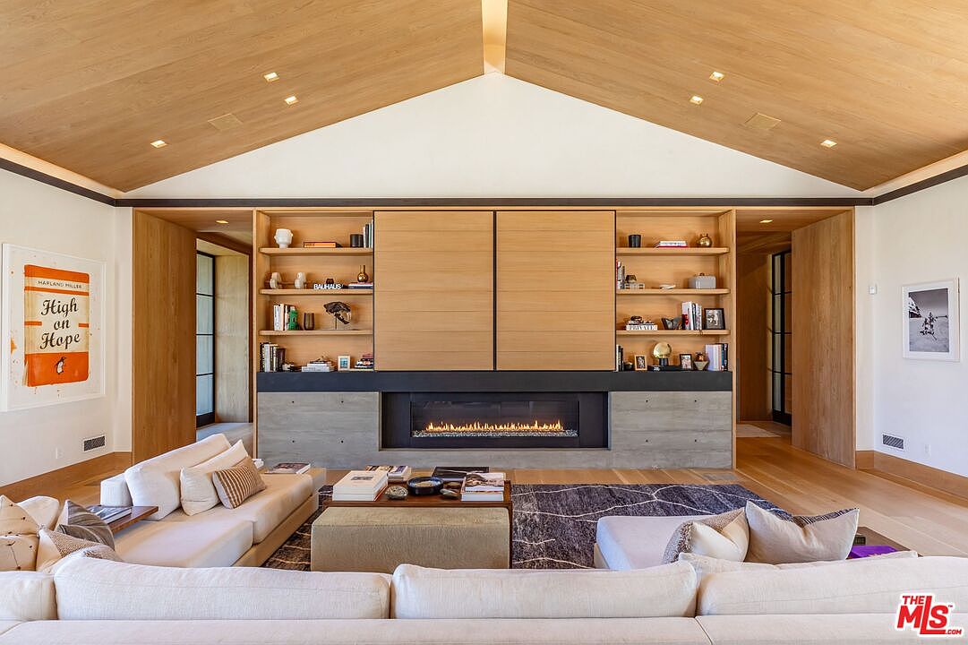 This sophisticated living room features a dramatic vaulted wood-paneled ceiling and a central fireplace integrated into a custom light-wood shelving unit. The space is anchored by a large, plush sectional sofa and a textured area rug, creating a warm and inviting atmosphere. The symmetrical design, combined with minimalist decor and clean lines, exudes a high-end, contemporary aesthetic perfect for luxury living.