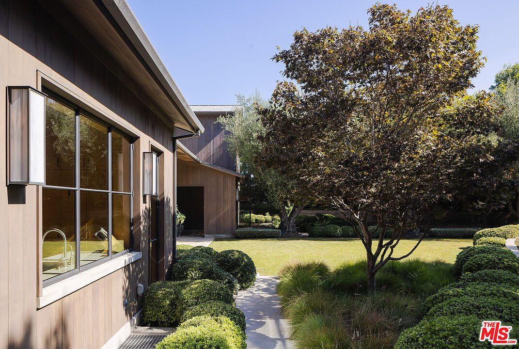 This side view of a modern residence showcases clean architectural lines with dark vertical wood siding and large, grid-paned windows. A manicured lawn and a lush, landscaped garden with neatly trimmed hedges and a mature tree create a serene, private atmosphere. The perspective captures the transition from the home's exterior to the expansive, well-maintained backyard space.