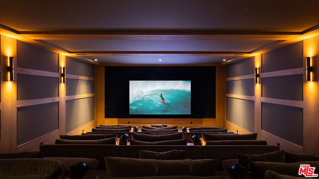 This image showcases a luxurious, tiered home theater room designed for an immersive cinematic experience. The space features plush, dark-toned seating arranged in rows facing a large, high-definition projection screen displaying a surfing scene. Warm, ambient wall sconces and recessed ceiling lighting create a sophisticated, cozy atmosphere perfect for private screenings.