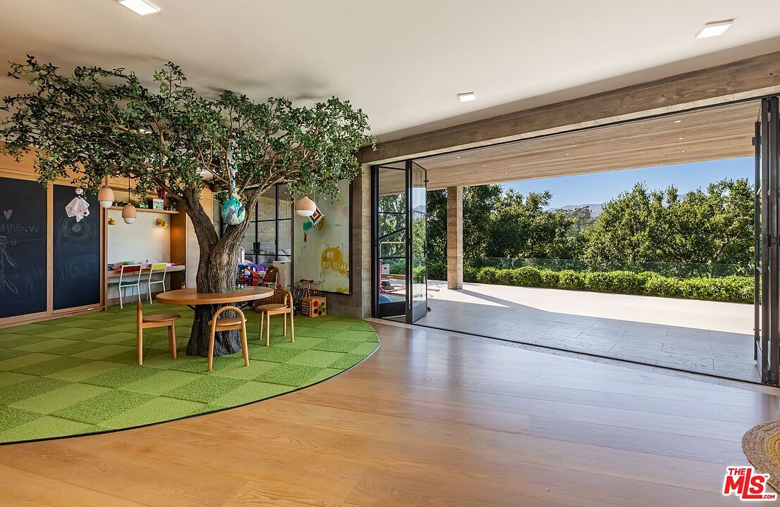 This unique interior space features a whimsical indoor tree integrated into a circular green rug, serving as a focal point for a children's play area. The room seamlessly transitions to a spacious outdoor patio through large, open glass doors, highlighting an indoor-outdoor living concept with views of lush greenery. The design combines natural elements with modern architecture, creating a bright, airy, and playful atmosphere.