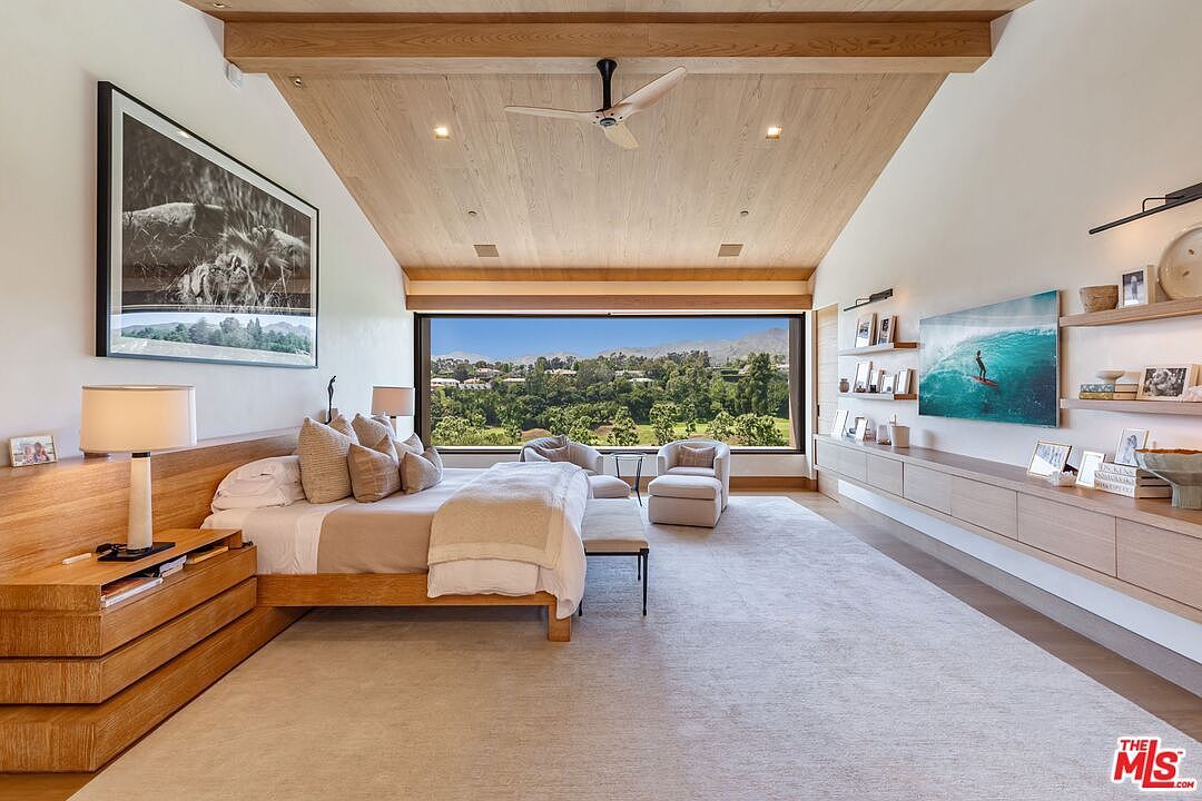 This luxurious primary bedroom features a vaulted wood-paneled ceiling, a large picture window framing a scenic landscape, and a custom built-in floating media console. The room is anchored by a platform bed with neutral-toned bedding, complemented by a soft area rug and minimalist, high-end wood cabinetry. The overall aesthetic is serene, modern, and sophisticated, emphasizing natural light and clean architectural lines.