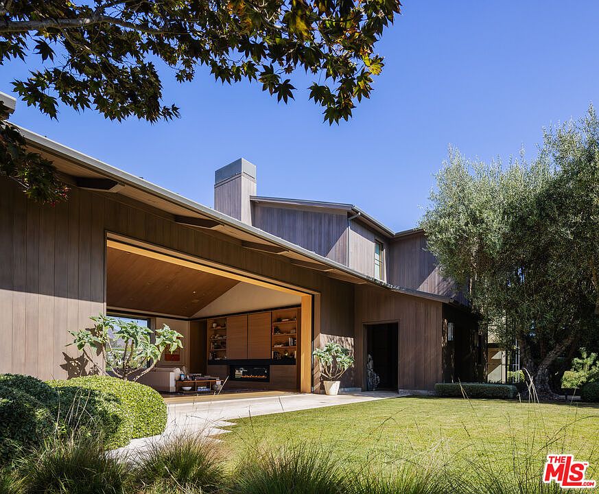 This modern architectural home features a seamless indoor-outdoor transition with a large, open-air living space that flows directly onto a manicured lawn. The exterior is clad in warm-toned vertical wood siding, complemented by a clean, minimalist design and a prominent chimney stack. The scene is bathed in bright, natural sunlight, highlighting the lush landscaping and the inviting, sophisticated atmosphere of the property.