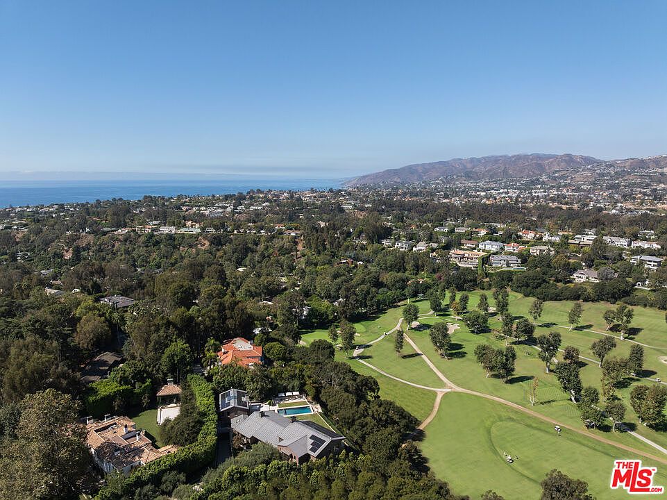 This high-angle aerial view captures a sprawling residential estate nestled within a lush, tree-filled landscape adjacent to a manicured golf course. The property features multiple structures, including a main residence with a dark roof and a secondary home with a red-tiled roof, both surrounded by dense greenery and private swimming pools. In the distance, the expansive Pacific Ocean meets the horizon, providing a stunning coastal backdrop that emphasizes the prime location and serene atmosphere of the estate.