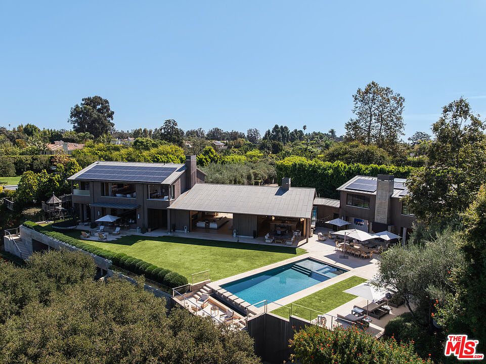 This stunning aerial perspective captures a sprawling, modern luxury estate nestled in a lush, green landscape. The property features a large, rectangular swimming pool, a manicured lawn, and an expansive open-air pavilion that seamlessly connects the indoor and outdoor living spaces. With its contemporary architectural design, solar panels, and serene surroundings, the home exudes an atmosphere of sophisticated, high-end living.