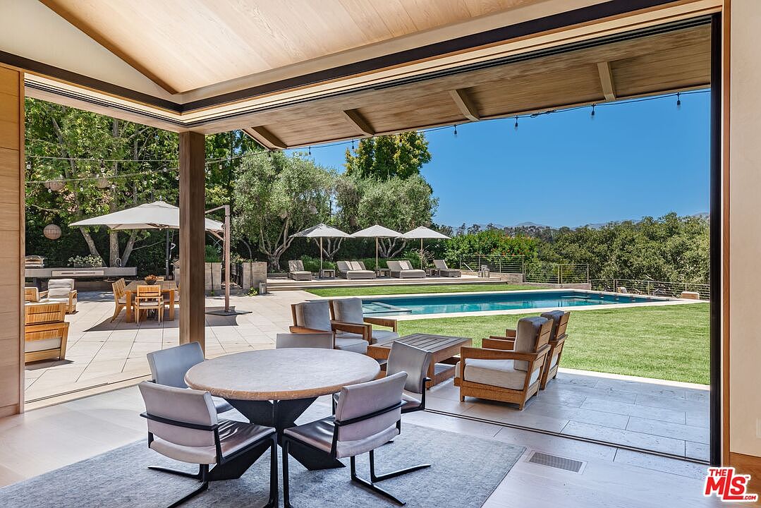 This image captures a seamless indoor-outdoor living space, featuring a covered patio area with a modern dining set that transitions into a sprawling backyard. The outdoor space includes a pristine swimming pool, multiple lounge chairs under umbrellas, and a well-manicured lawn, all set against a backdrop of lush trees and distant mountains. The perspective is from inside looking out, emphasizing the expansive, open-concept design and the luxurious, resort-like atmosphere of the property.
