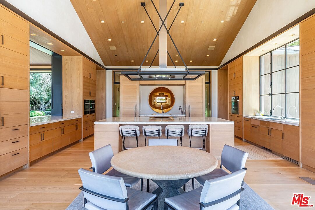 This expansive, high-end kitchen features warm wood cabinetry, a large central island with bar seating, and a dedicated circular dining area in the foreground. The room is defined by its vaulted wood-paneled ceiling, a striking modern light fixture, and a symmetrical layout that emphasizes luxury and clean, contemporary design. Large windows and integrated appliances create a seamless, sophisticated space perfect for both culinary tasks and social gatherings.
