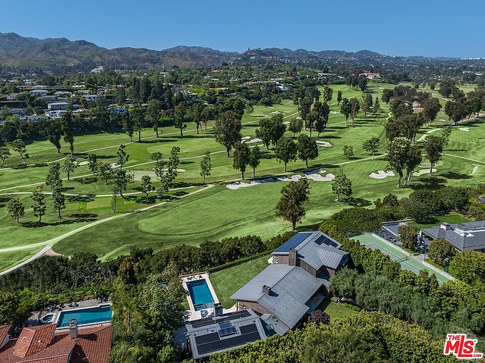 This high-angle aerial shot captures a luxurious estate property overlooking a sprawling, lush green golf course set against a backdrop of rolling hills. The home features a modern architectural design with dark metal roofing, solar panels, and two distinct swimming pools nestled within private, landscaped grounds. The perspective emphasizes the property's prime location, offering a seamless blend of upscale residential living and expansive outdoor recreation.