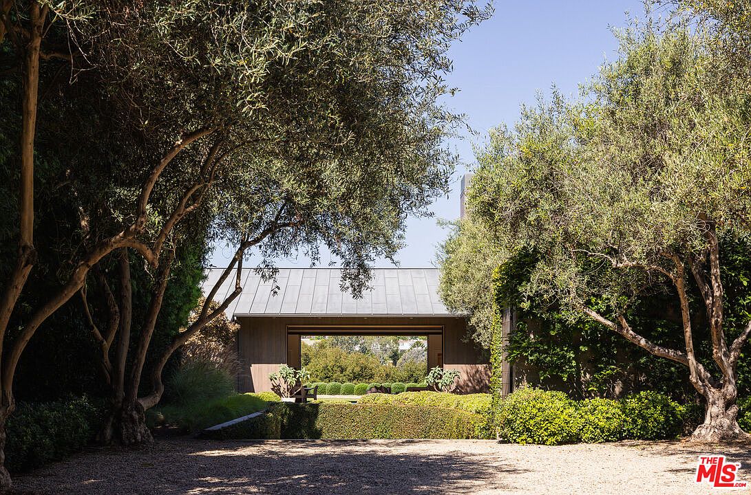 This serene outdoor scene features a modern, minimalist structure with a metal roof that frames a view through to the lush greenery beyond. The foreground is composed of a gravel driveway flanked by mature, sculptural olive trees and manicured hedges, creating a sophisticated and tranquil atmosphere. The perspective is eye-level, emphasizing the seamless integration between the architectural design and the surrounding natural landscape.