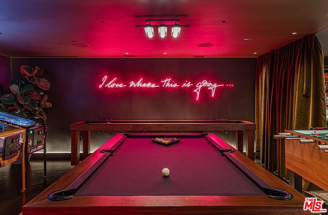 This vibrant game room features a central pool table with a purple felt surface, set against a dark wall adorned with a glowing neon sign that reads 'I love where this is going...'. The space is equipped with a Star Wars pinball machine on the left and a foosball table on the right, creating an inviting entertainment area. The room is illuminated by a modern industrial-style ceiling light fixture and warm accent lighting, giving it a sophisticated yet playful atmosphere.