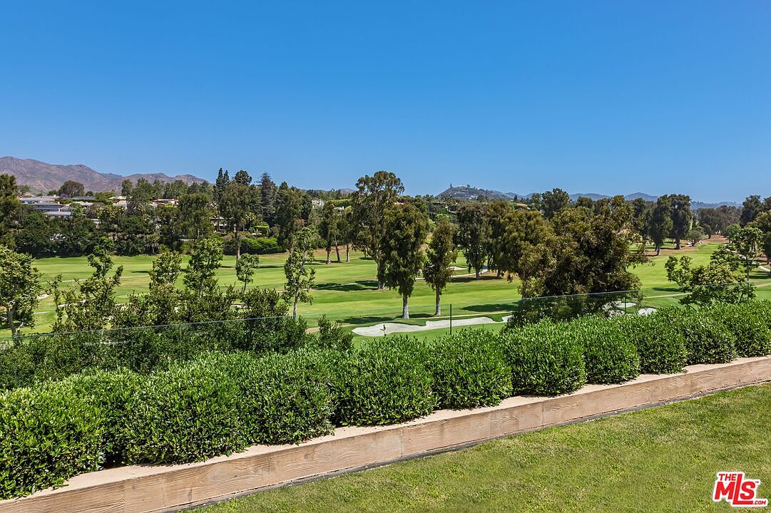 This elevated perspective captures a scenic view of a lush green golf course from a private balcony or terrace. The foreground features a neatly manicured hedge and a wooden retaining wall, while the background showcases rolling hills and mature trees under a clear blue sky. The scene conveys a sense of tranquility and luxury, highlighting the property's prime location overlooking expansive outdoor recreational space.