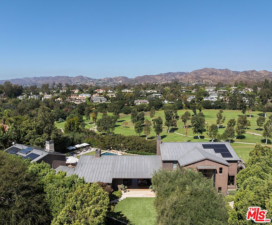 This high-angle aerial shot captures a sprawling, modern estate nestled against a lush green golf course and rolling hills. The home features a distinctive dark metal roof, solar panels, and a seamless blend of indoor-outdoor living spaces, including a private backyard lawn and a hidden pool area. The expansive landscape and clear blue sky create a serene, luxurious atmosphere perfect for a high-end property listing.