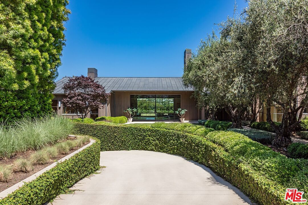 This elegant, modern estate features a clean-lined architectural design with a metal roof and warm-toned wood siding. A manicured, curved hedge lines the concrete driveway, leading the eye toward a large, floor-to-ceiling glass entryway that reflects the surrounding lush greenery. The scene is framed by mature trees and vibrant landscaping, creating a serene and sophisticated atmosphere.