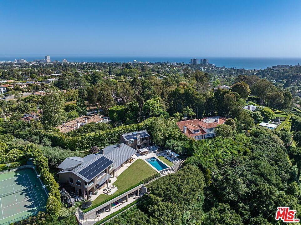 This high-angle aerial view captures a sprawling luxury estate nestled within a lush, tree-filled landscape overlooking the distant coastline. The property features a modern multi-level residence with solar panels, a private tennis court, and a pristine swimming pool set against a backdrop of dense greenery. The perspective emphasizes the expansive nature of the grounds and the seamless integration of the home into its scenic, elevated environment.