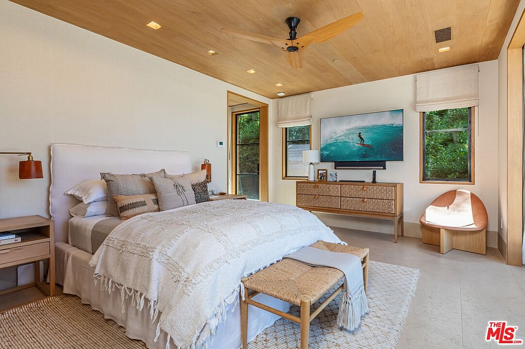 This serene primary bedroom features a warm, organic aesthetic with a light wood-paneled ceiling, a textured white bedspread, and a woven bench at the foot of the bed. The room is bathed in natural light from large windows and a glass door, highlighting the neutral color palette and modern, minimalist furniture. A large flat-screen television mounted above a wooden dresser serves as a focal point, creating a sophisticated and relaxing retreat.