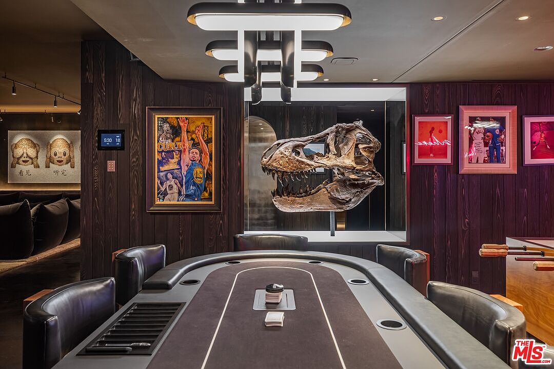 This sophisticated game room features a custom poker table as the centerpiece, surrounded by sleek, dark leather chairs. The room is accented by dark wood-paneled walls, a striking T-Rex skull display case, and curated sports-themed artwork, creating a masculine and high-end atmosphere. A modern, multi-tiered chandelier hangs above, casting a warm glow over the space, which also includes a foosball table to the side.