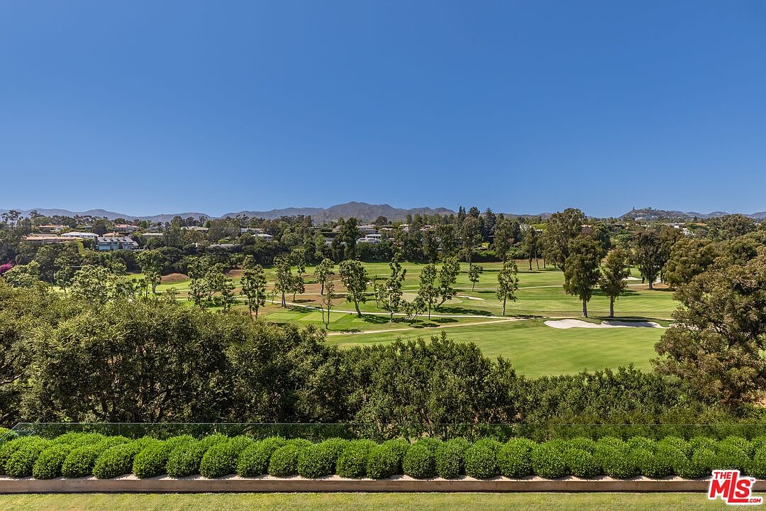 This elevated perspective captures a stunning, panoramic view of a lush green golf course from what appears to be a private balcony or terrace. In the foreground, a neatly manicured row of rounded hedges provides a structured border, while the background showcases rolling hills and a clear blue sky. The scene conveys a sense of tranquility, luxury, and expansive outdoor living space.