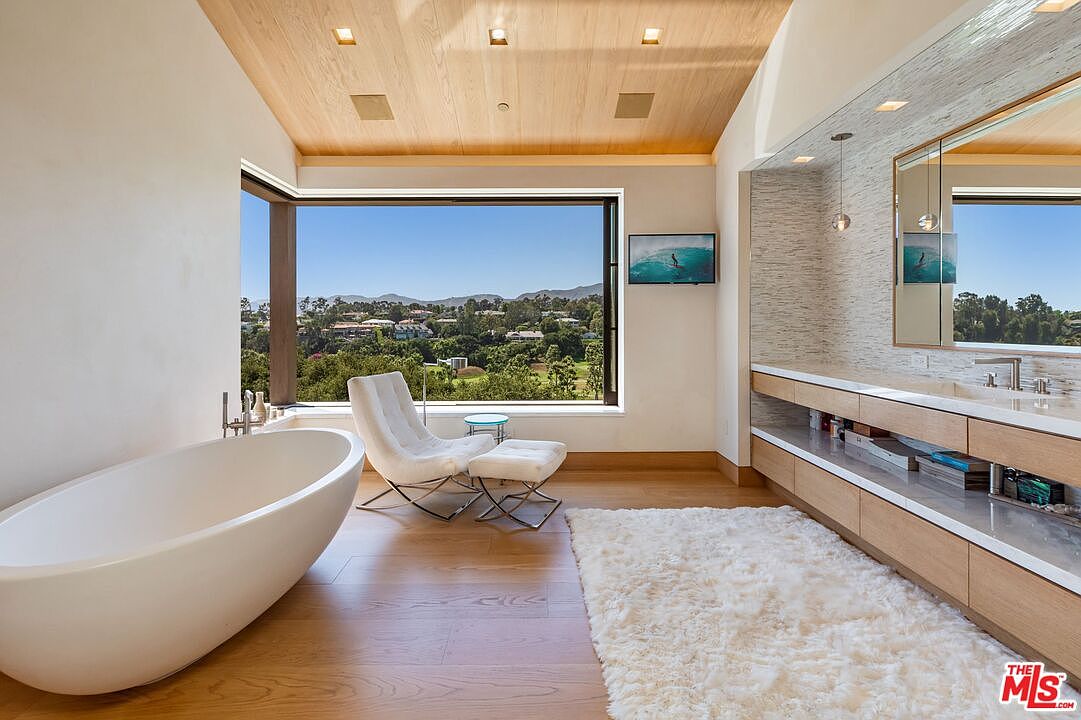 This luxurious primary bathroom features a sleek, freestanding soaking tub positioned to take advantage of expansive floor-to-ceiling windows with panoramic hillside views. The space is anchored by a long, custom wood vanity with a textured stone backsplash and a plush white area rug, creating a serene, spa-like atmosphere. The combination of warm wood ceilings, light hardwood floors, and modern minimalist design elements exudes high-end sophistication.