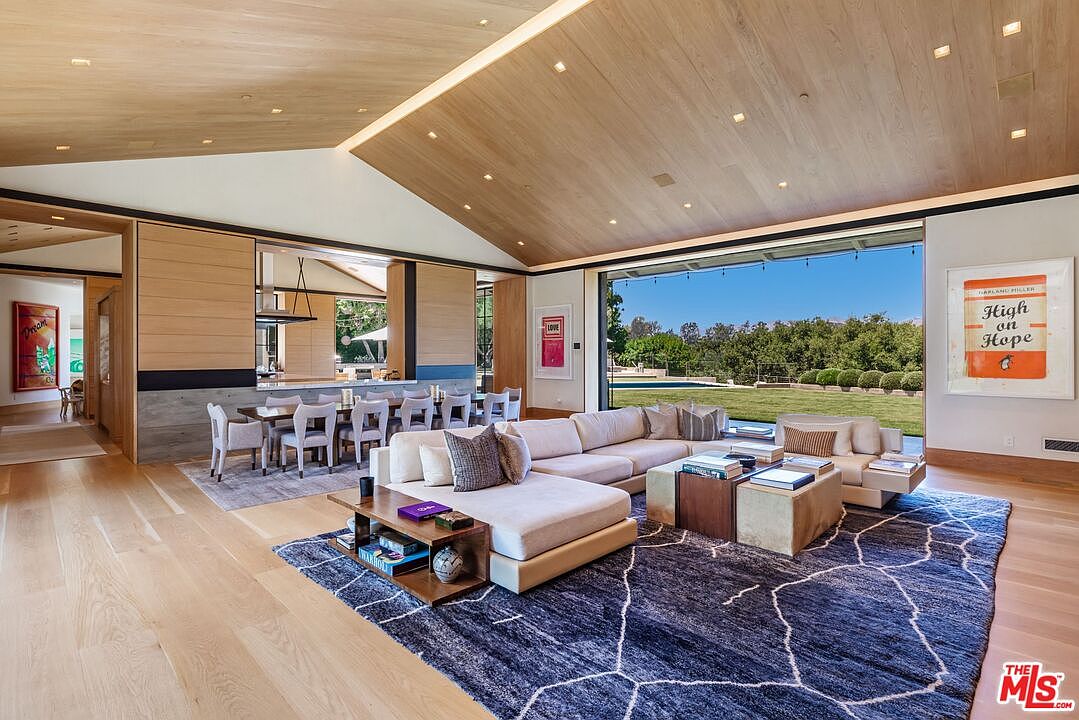 This expansive, open-concept living area features a vaulted wood-paneled ceiling and a seamless transition to the outdoors through a massive floor-to-ceiling glass wall. The space is anchored by a large, plush sectional sofa atop a bold, patterned navy blue rug, complemented by a contemporary dining area in the background. The interior design blends modern luxury with artistic flair, highlighted by vibrant wall art and warm, natural wood tones that create an inviting and sophisticated atmosphere.