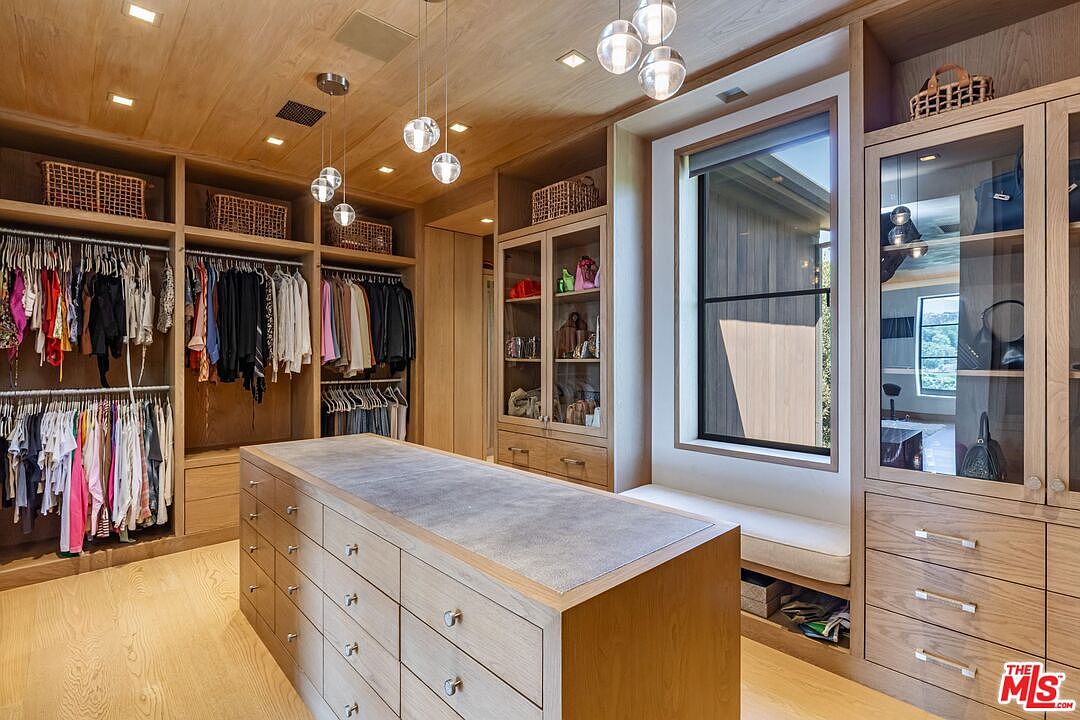 This luxurious walk-in closet features custom light-wood cabinetry, extensive hanging space, and a large central island with a grey stone countertop. The room is illuminated by modern glass pendant lights and recessed lighting, with a built-in window seat providing a cozy touch. The overall aesthetic is sophisticated, organized, and high-end, showcasing ample storage and premium finishes.