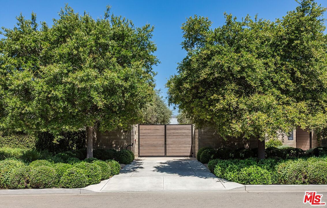 This image captures the private, gated entrance to a property, framed by two large, mature trees that provide a sense of seclusion and natural beauty. A modern, horizontal-slat wooden gate serves as the focal point, set against a clean concrete wall and a paved driveway. The scene conveys a sophisticated, minimalist aesthetic with lush, manicured landscaping that enhances the home's curb appeal.