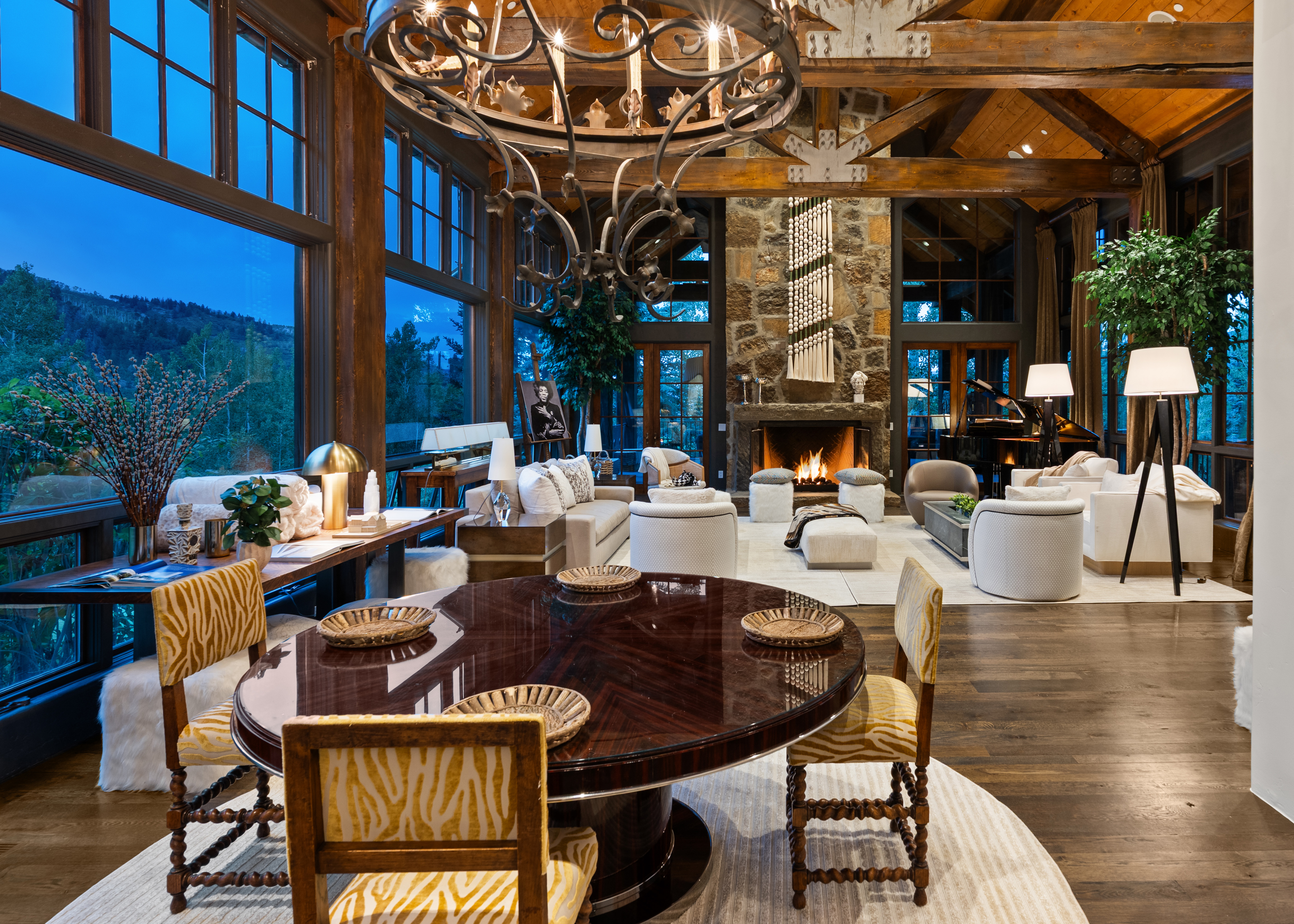 This is a luxurious living room featuring a stone fireplace, exposed wooden beams, and large windows offering a scenic view. The room is furnished with comfortable seating arrangements, including sofas and armchairs, arranged around a central coffee table and ottoman. A grand chandelier hangs above a dining table, adding to the room's elegance and grandeur.