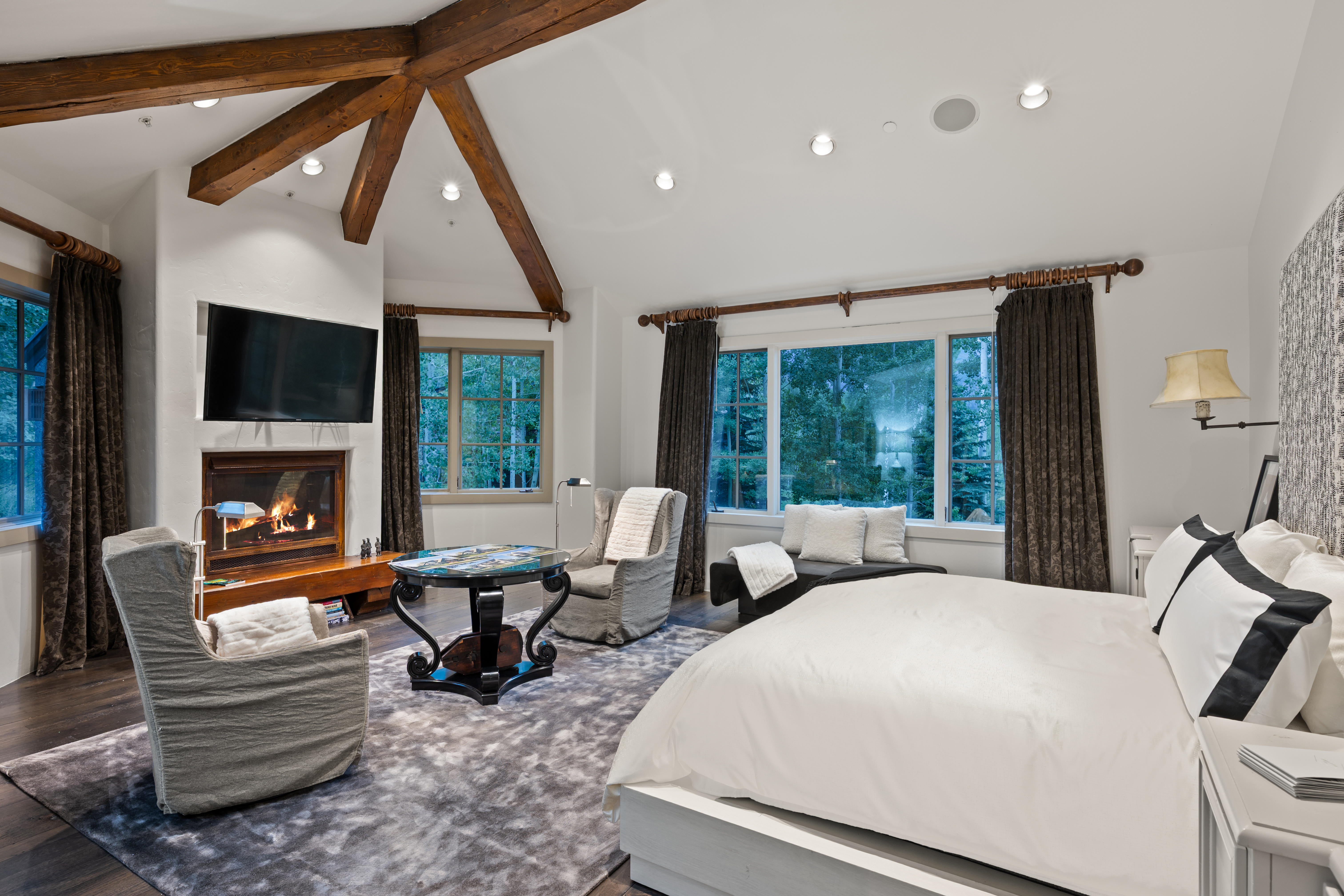 This is a luxurious primary bedroom featuring a king-size bed with white linens and black accents, two comfortable armchairs, and a fireplace with a mounted television. The room has exposed wooden beams, recessed lighting, and large windows offering a view of the surrounding trees. The overall style is elegant and cozy, creating a relaxing retreat.