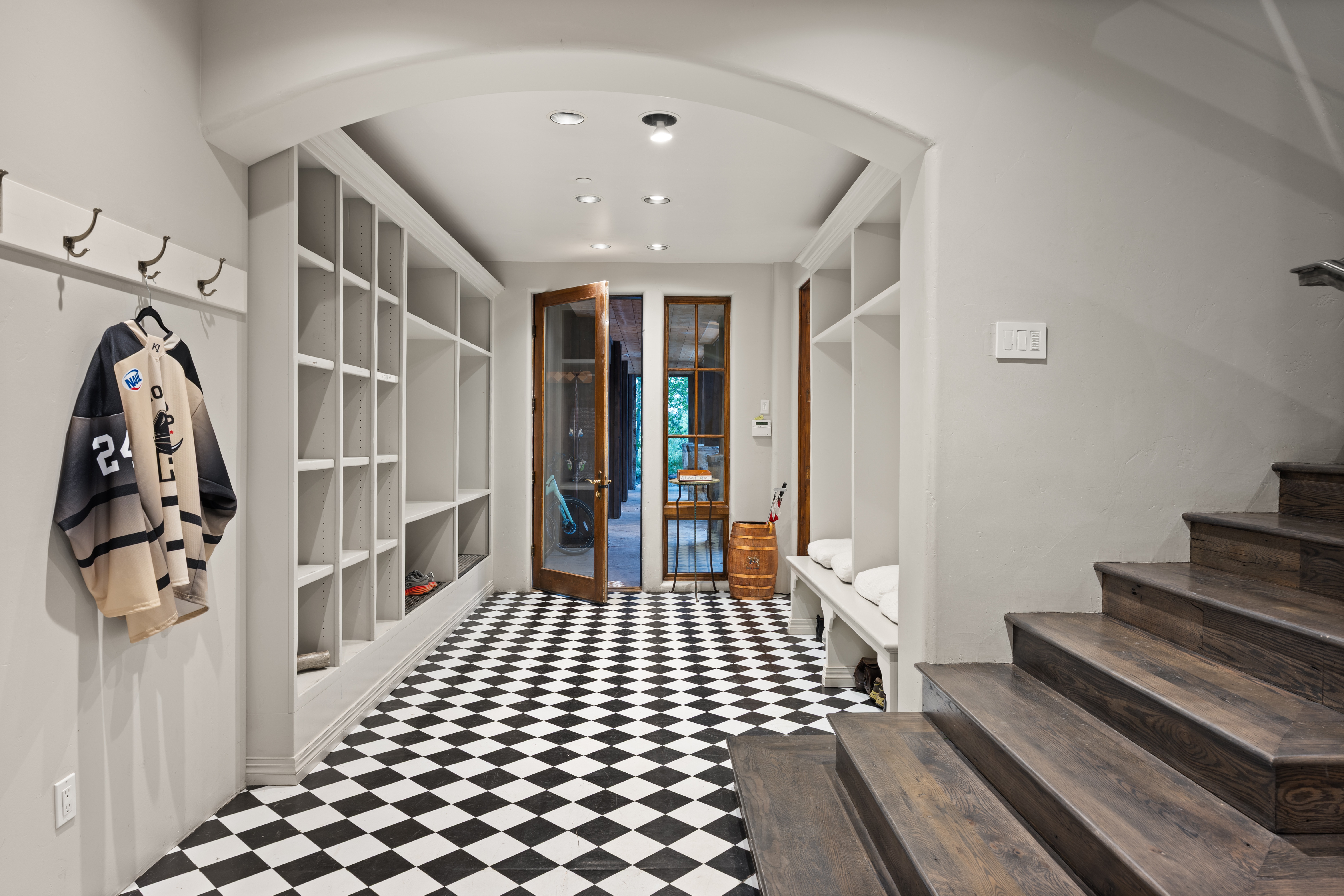 This interior shot showcases a stylish hallway with a striking black and white checkered floor. Built-in shelving units line the walls, providing ample storage, while a doorway leads to an outdoor area. A wooden staircase adds a touch of warmth and elegance to the space.