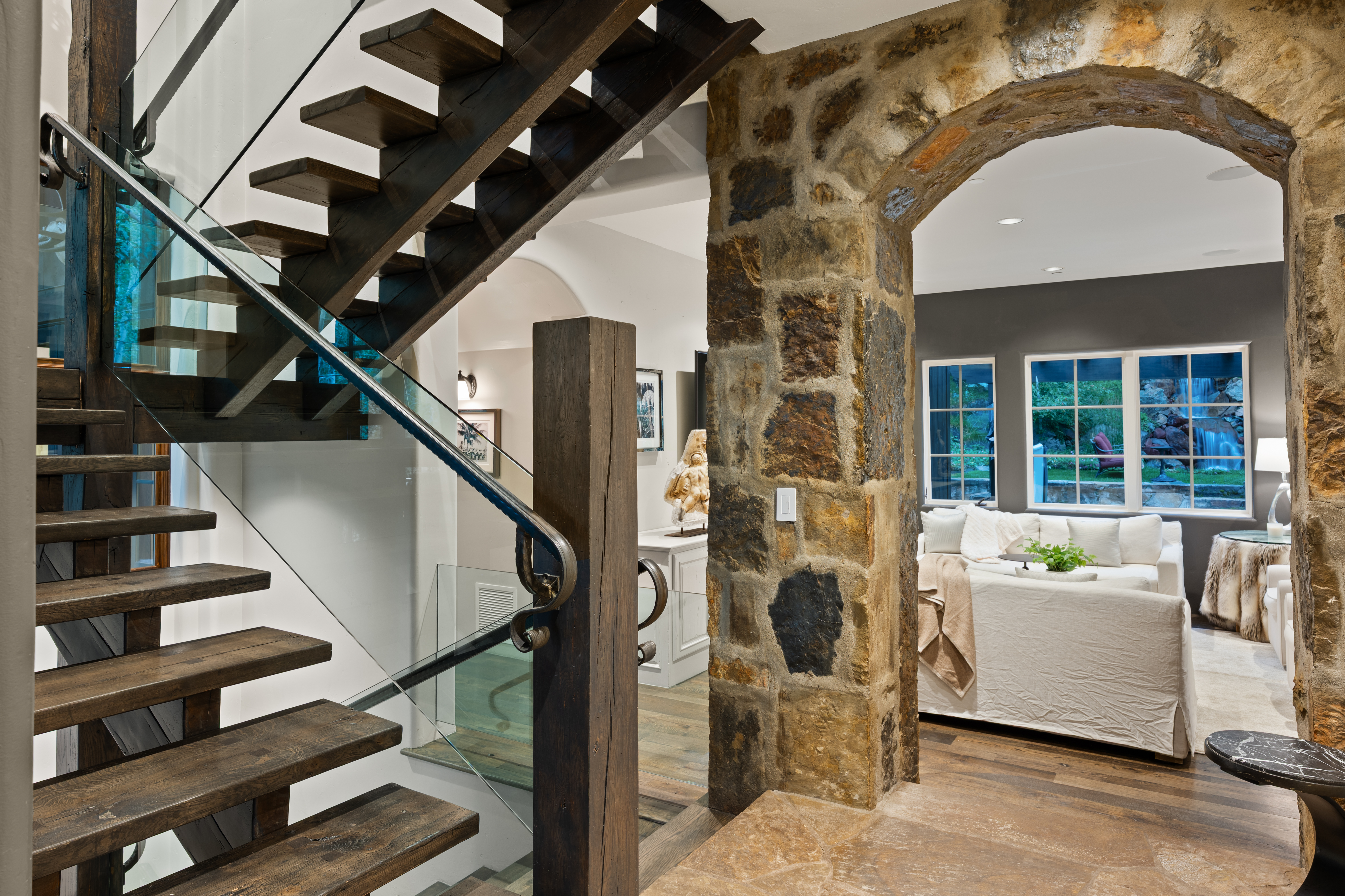 This interior shot showcases a unique hallway with striking architectural details. A rustic wooden staircase with glass railings ascends, while a stone archway leads into a living area with a white sofa and natural light streaming through the windows. The combination of wood, stone, and glass creates a sophisticated and inviting atmosphere.