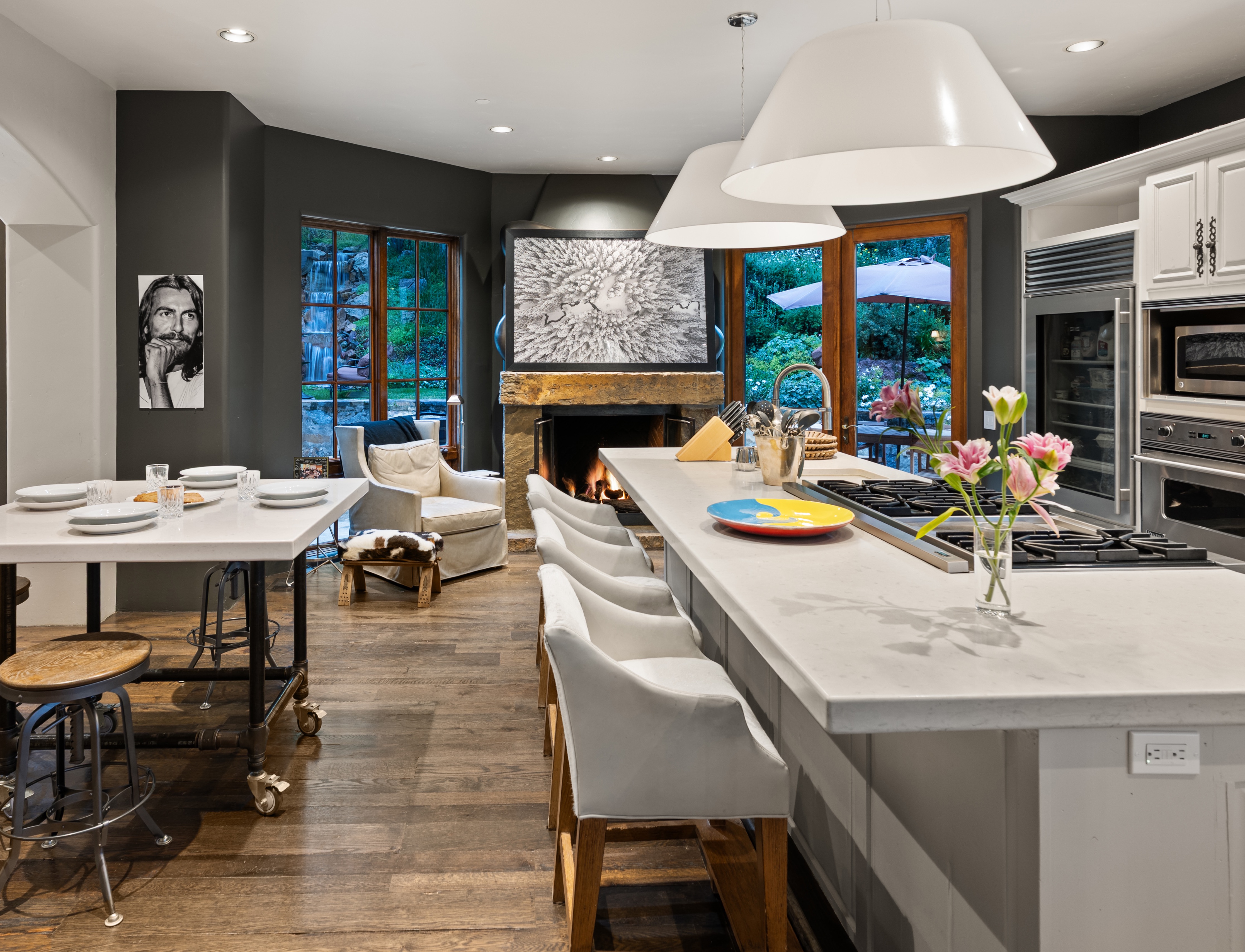 This is a well-lit kitchen featuring a large island with a white countertop and white chairs. The kitchen has a fireplace and a dining table with a unique design. The overall style is modern and luxurious, with a focus on natural light and open space.