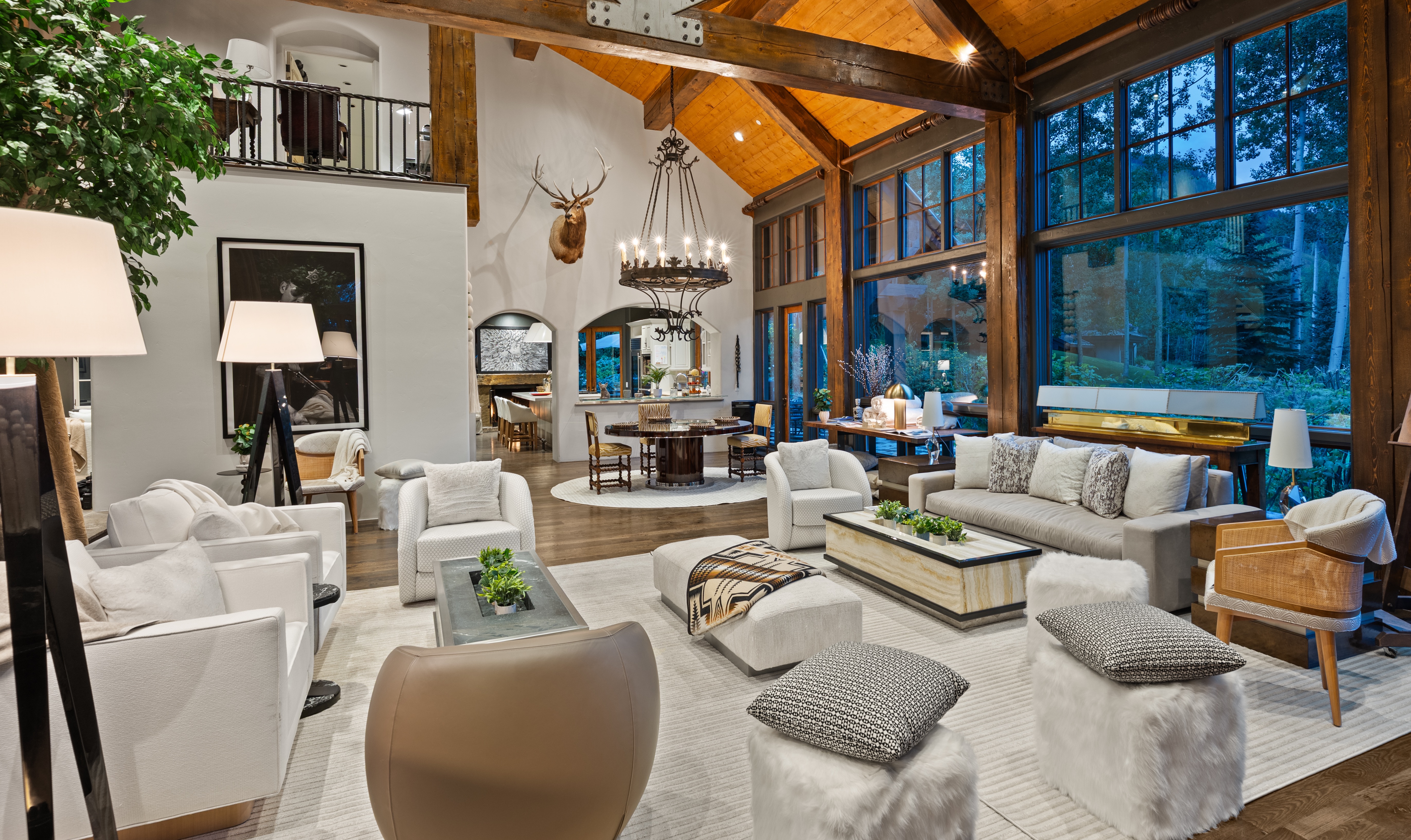 This is an interior shot of a luxurious living room featuring a high vaulted ceiling with exposed wooden beams and large windows offering natural light. The room is furnished with white sofas and chairs, a unique coffee table with greenery, and decorative ottomans, all arranged on a light-colored rug. A mounted deer head adds a rustic touch, while a chandelier hangs above a dining area visible in the background, creating an open and inviting space.