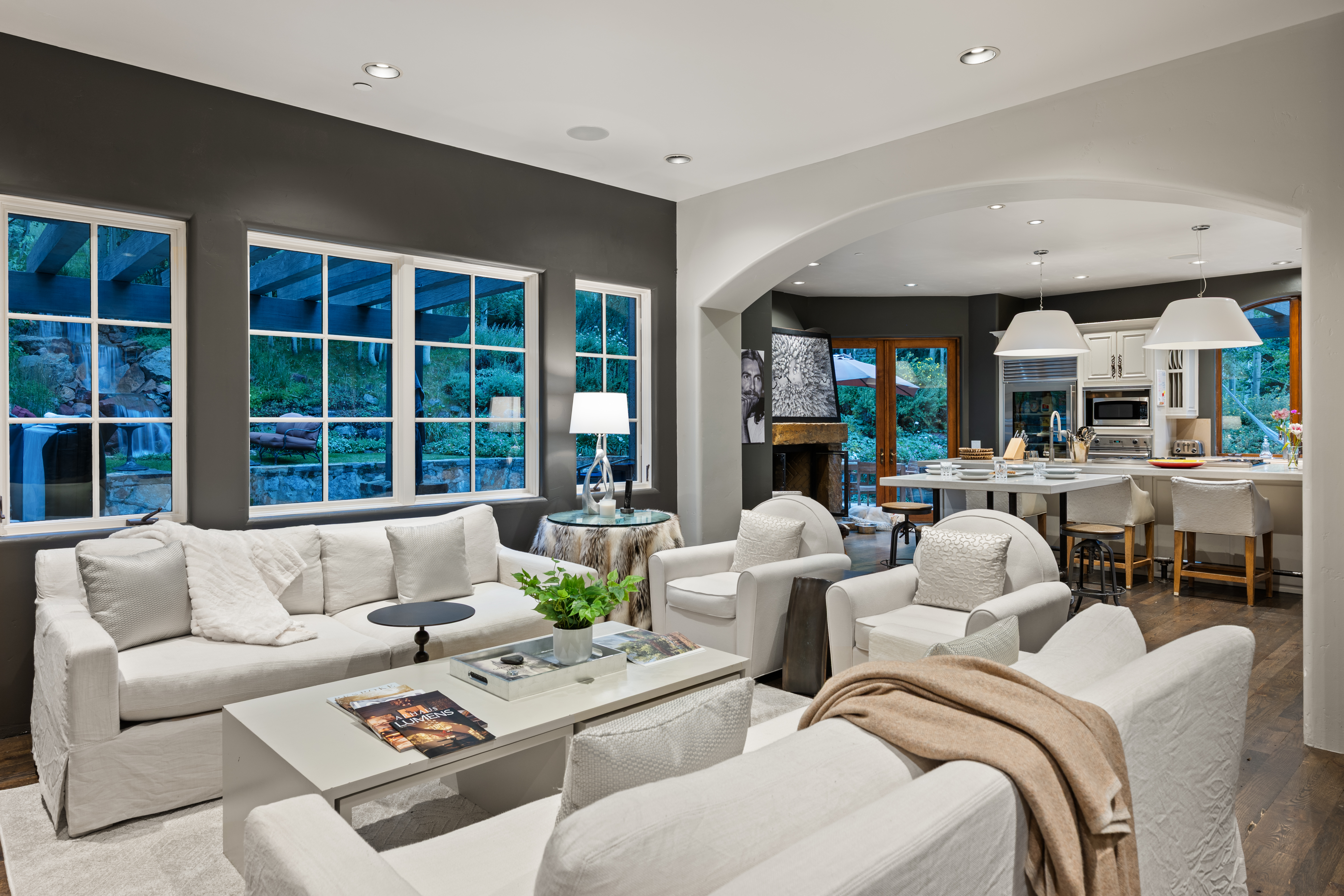 This is an interior shot of a well-lit living room that seamlessly transitions into a kitchen area. The living room features a neutral color palette with white sofas and armchairs, a light gray coffee table, and dark gray accent walls. Large windows offer a view of the outdoor landscape, and an arched opening leads to the kitchen, which includes a kitchen island with bar seating and pendant lighting. The overall impression is one of spaciousness and modern elegance.