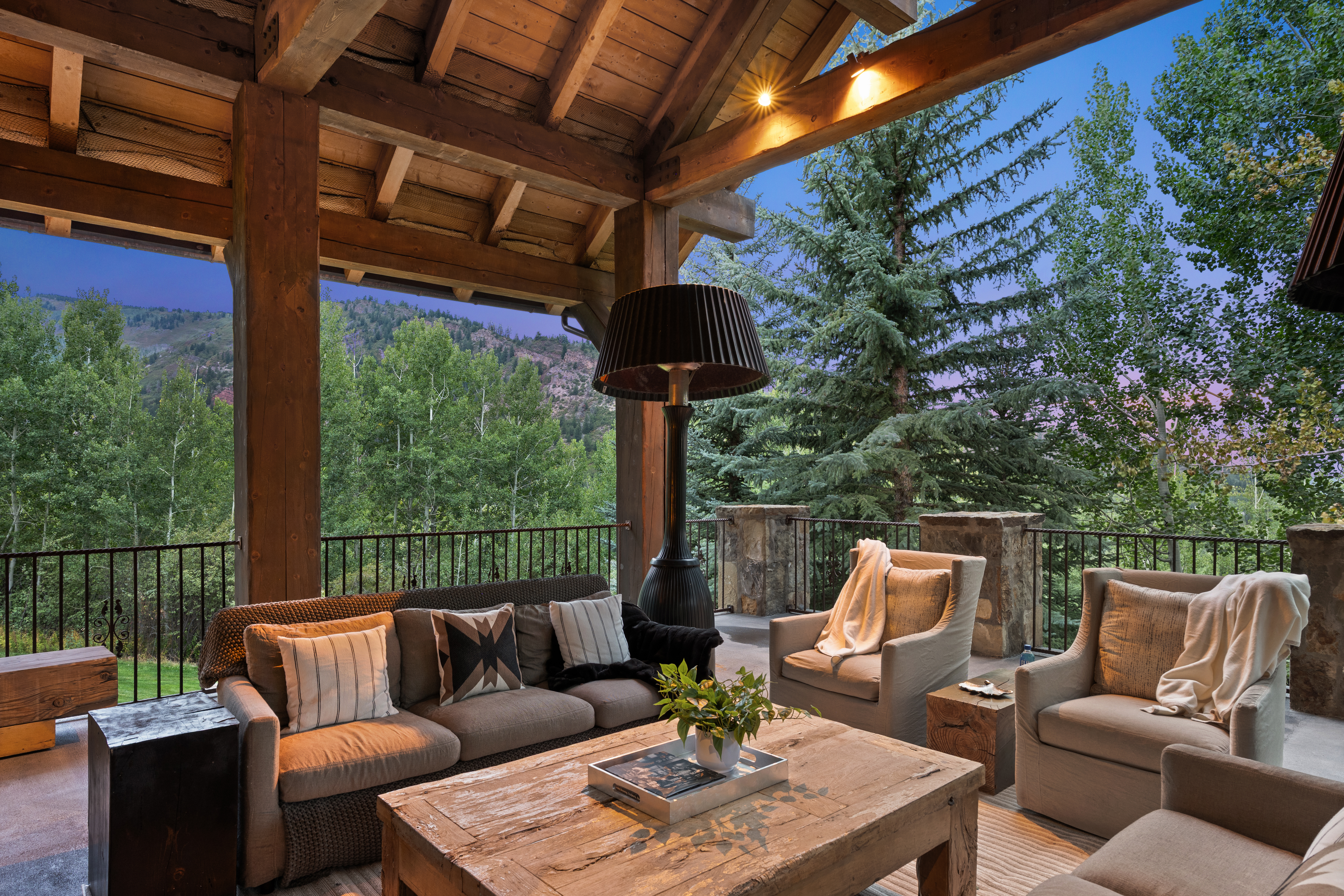 This image showcases a beautifully furnished outdoor patio or balcony area. The space features comfortable seating arrangements, including a sofa and armchairs, arranged around a rustic wooden coffee table. The patio is covered by a wooden roof structure with exposed beams, and offers a scenic view of trees and mountains in the background, creating a relaxing and inviting atmosphere.