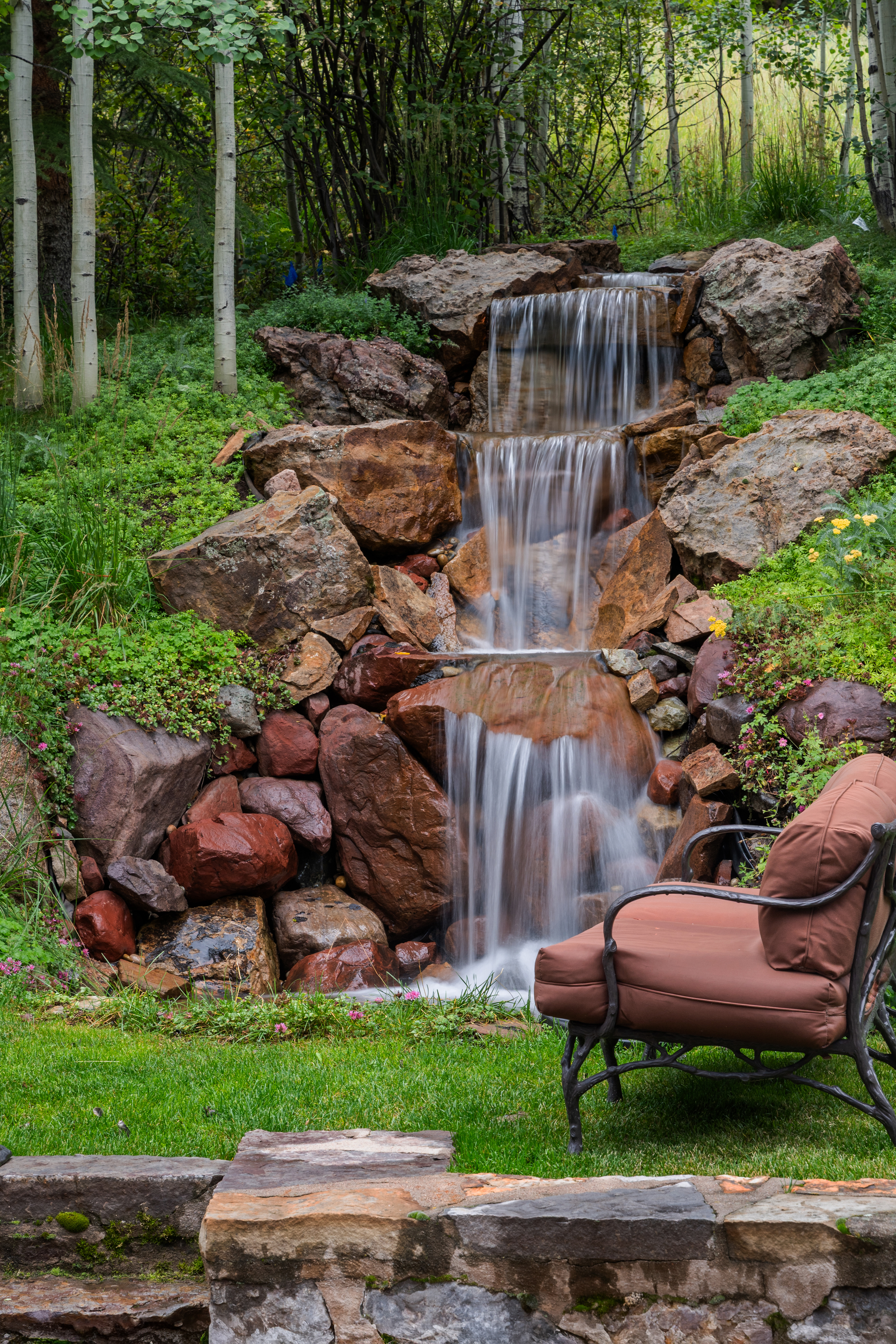 This image showcases a serene backyard garden featuring a multi-tiered rock waterfall as its focal point. Lush greenery and mature trees surround the waterfall, creating a tranquil and private outdoor space. A comfortable chair is placed nearby, inviting relaxation and enjoyment of the natural setting.