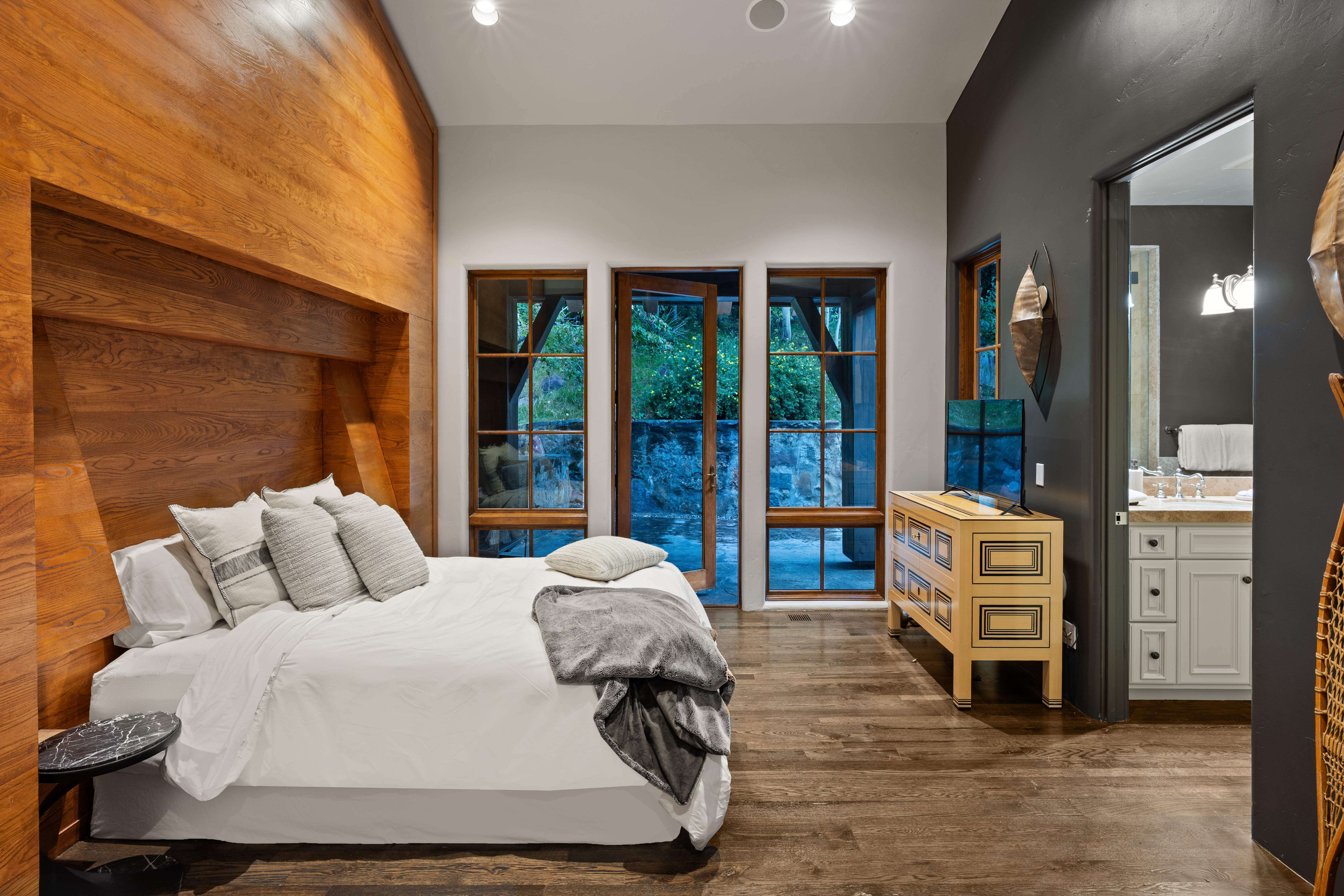 This is a primary bedroom featuring a wooden accent wall behind the bed, which has white linens and gray pillows. Three large windows provide natural light and a view of the outdoors. A wooden dresser with a television sits to the side, and a glimpse into the bathroom is visible, creating a luxurious and inviting space.