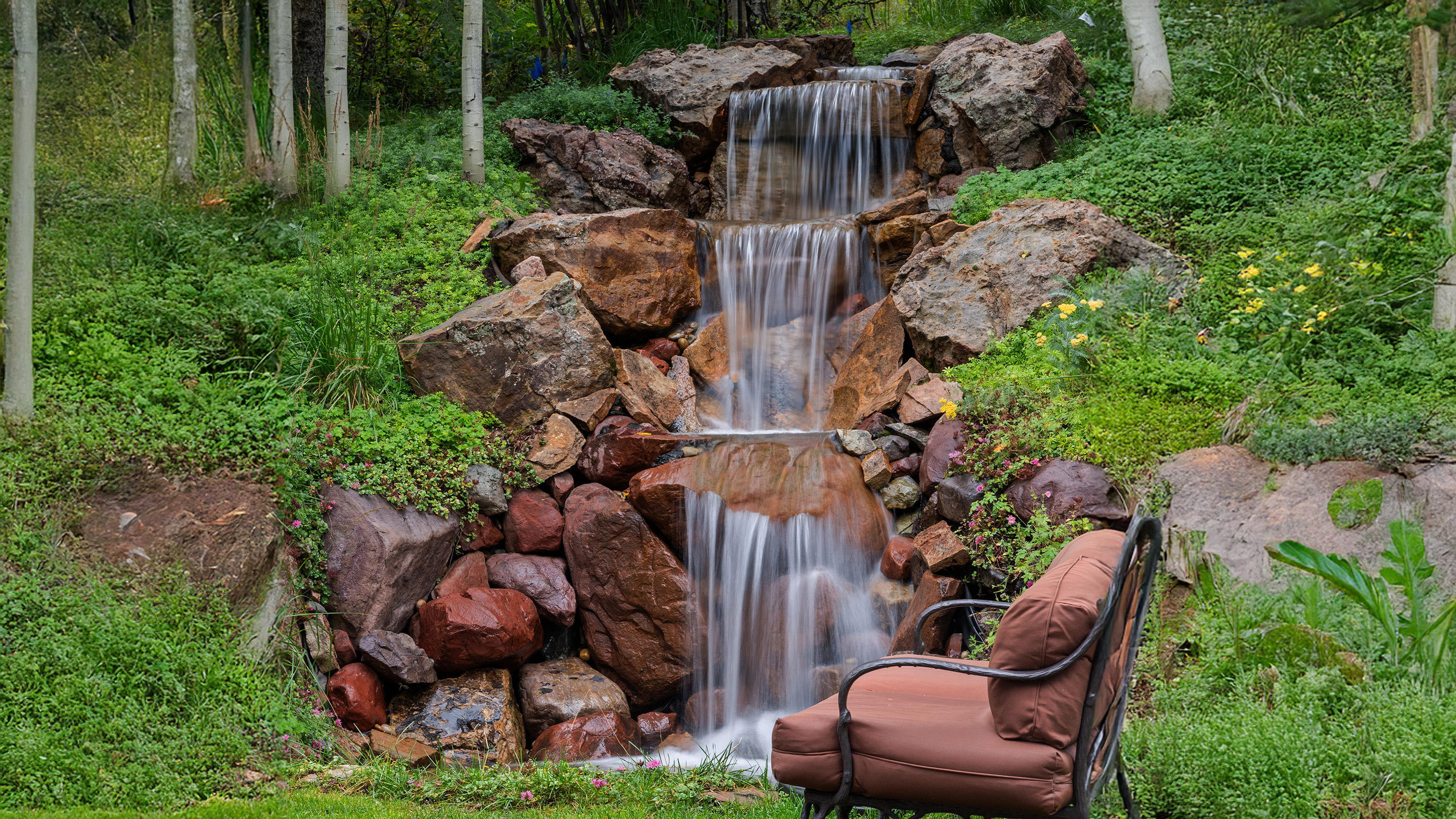 This image showcases a tranquil garden featuring a multi-tiered waterfall cascading over natural rocks, enhancing the property's appeal with serene water sounds and visual interest. Lush greenery surrounds the waterfall, creating a private and peaceful outdoor setting, while a comfortable chair offers a place to relax and enjoy the scenery. The scene suggests a well-maintained and inviting backyard oasis.