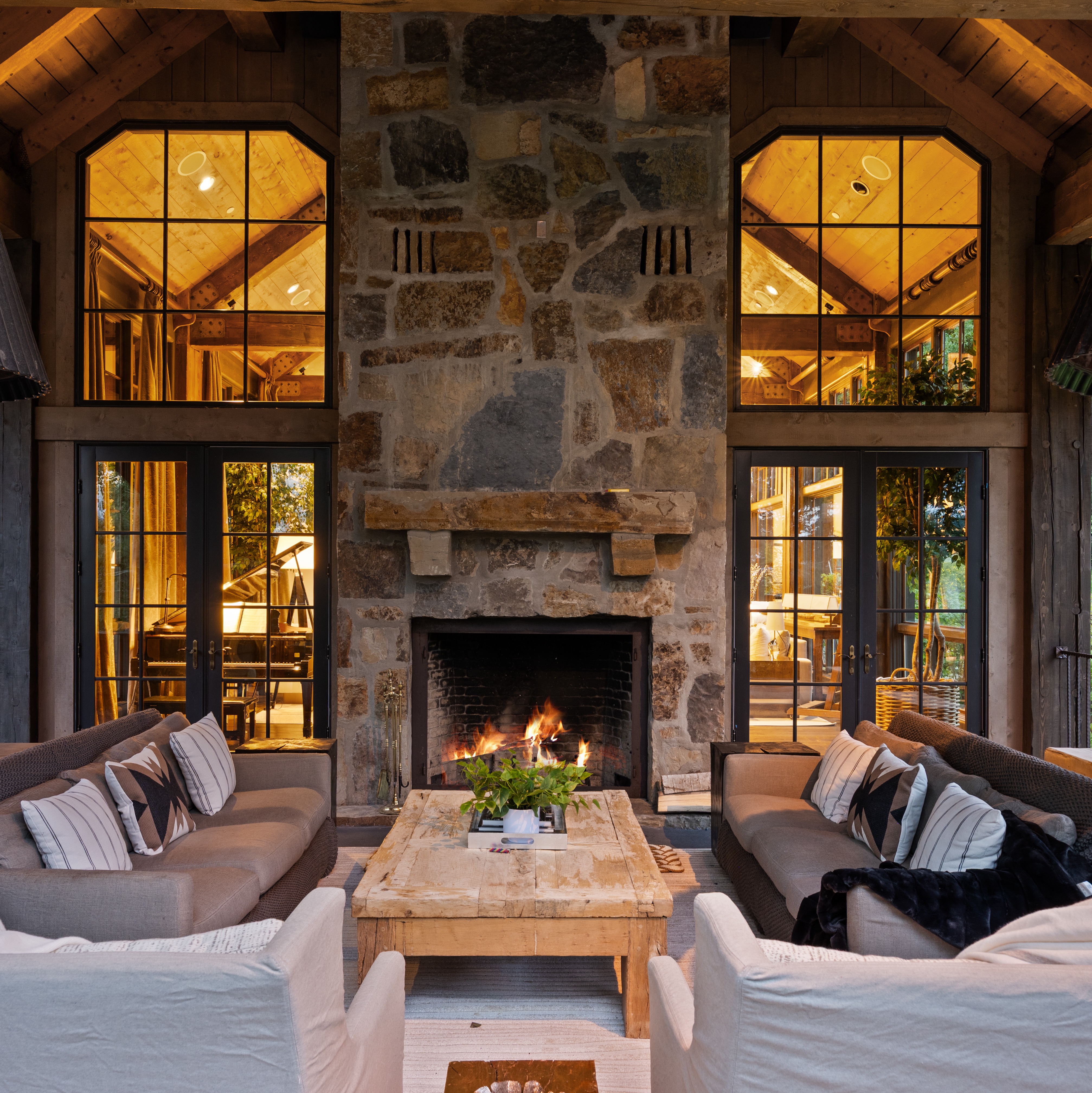 This inviting living room features a grand stone fireplace as its focal point, flanked by large windows and doors that offer views of the surrounding landscape. Two comfortable sofas and armchairs are arranged around a rustic wooden coffee table, creating a cozy and sophisticated atmosphere. The room's design blends natural materials with elegant furnishings, perfect for relaxation and entertaining.