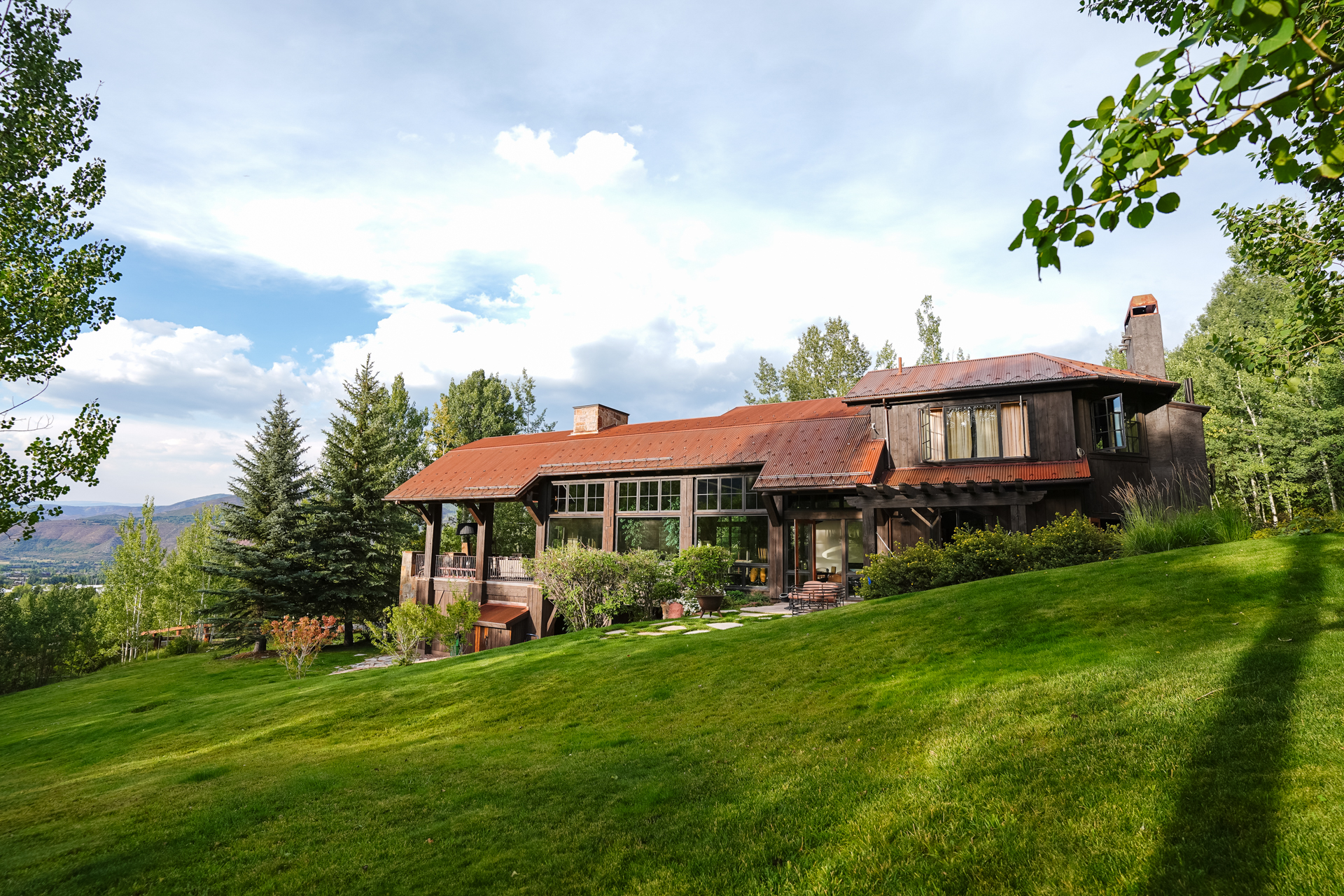 This image showcases the side view of a luxurious home nestled on a lush green hillside. The house features a distinctive rusty metal roof, dark wood siding, and numerous windows that offer stunning views. The surrounding landscape includes mature trees and manicured lawns, creating a serene and private setting.