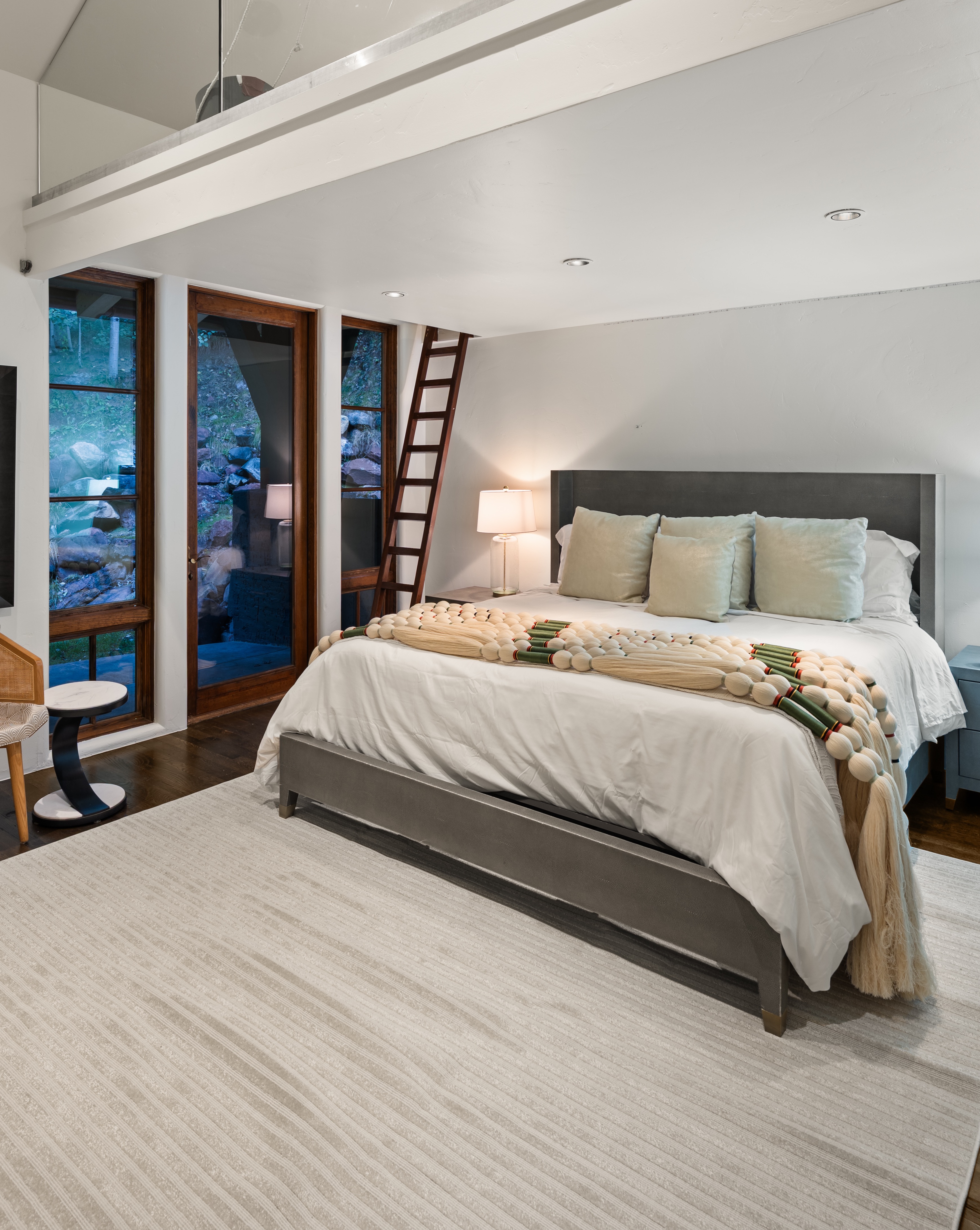 This is a primary bedroom featuring a gray upholstered bed with light-colored pillows and a decorative throw. The room has large windows offering a view of the outdoors, and a wooden ladder leads to an upper level. The flooring is dark wood, complemented by a light-colored area rug, creating a serene and inviting atmosphere.
