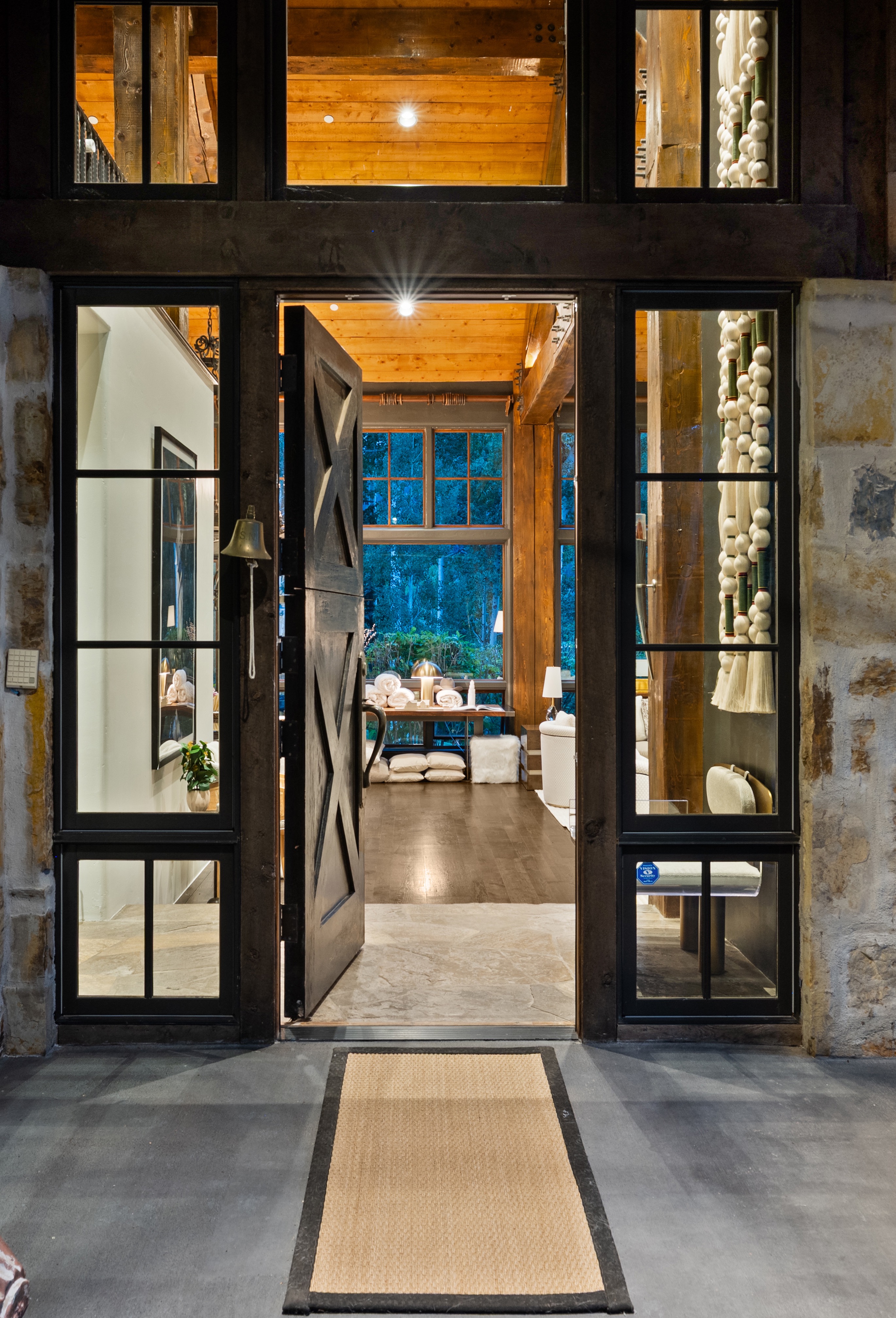 This image showcases an inviting entryway with a rustic yet elegant design. The open wooden door, flanked by large windows, reveals a glimpse of the interior, featuring wooden beams and stylish decor. A woven doormat adds a touch of warmth to the stone flooring, creating a welcoming first impression.