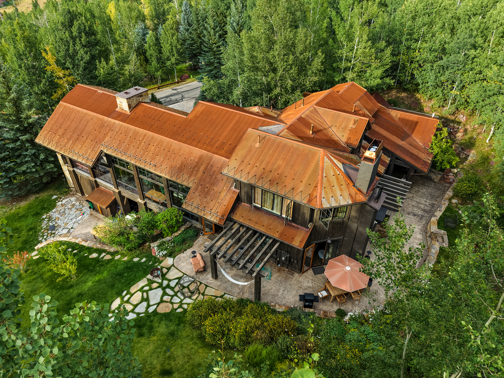 This aerial shot showcases a large, rustic-style home nestled among lush greenery. The house features a distinctive rusted metal roof, dark wood siding, and multiple outdoor living spaces, including a patio with a pergola, seating, and a grill. Stone pathways meander through the well-maintained yard, creating an inviting and private retreat.