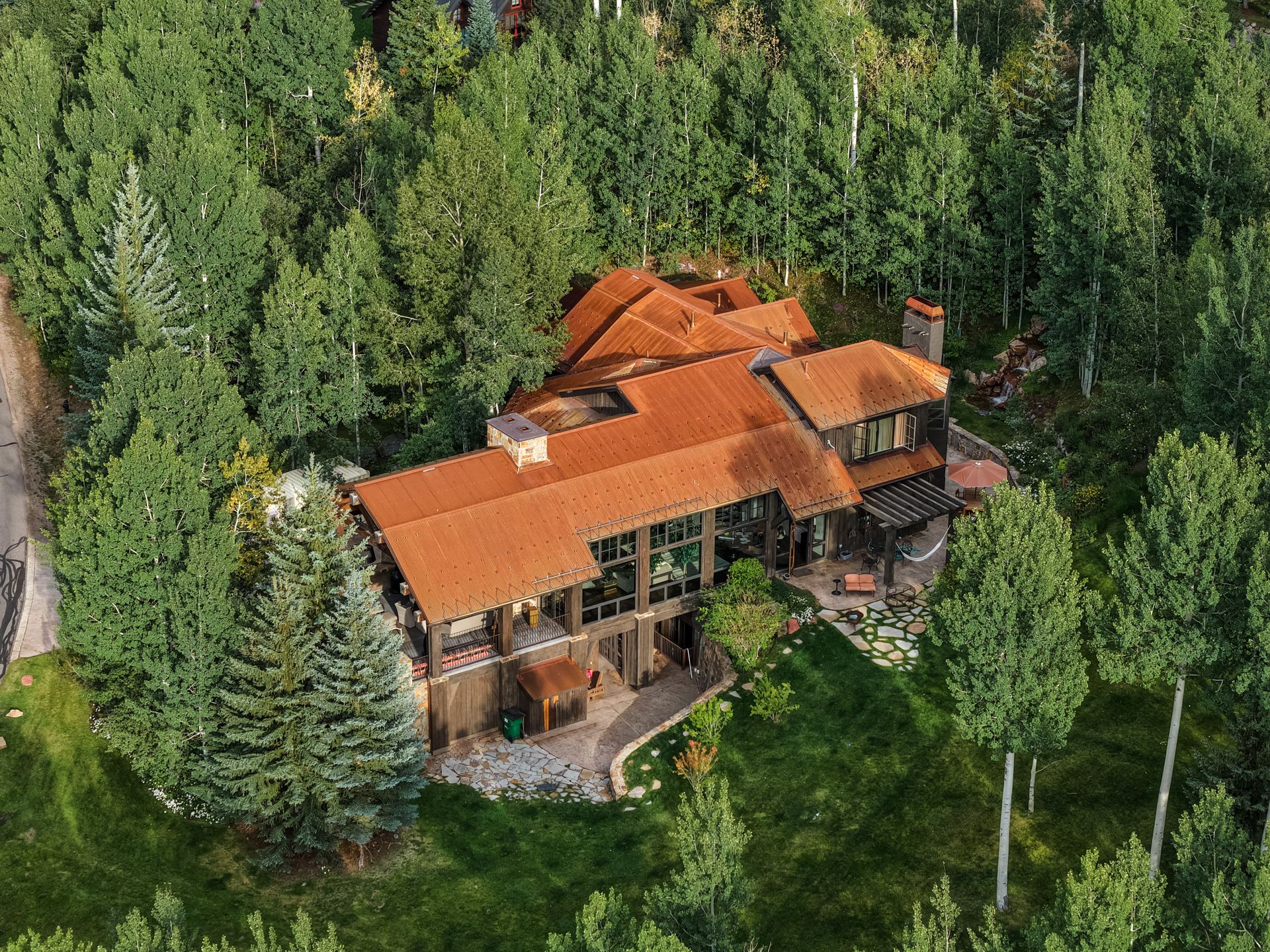 This aerial view showcases a luxurious multi-story home nestled among lush green trees. The house features a distinctive rusted metal roof, large windows, and a stone patio area with outdoor seating. The surrounding landscape includes well-maintained lawns and mature trees, creating a sense of privacy and tranquility.