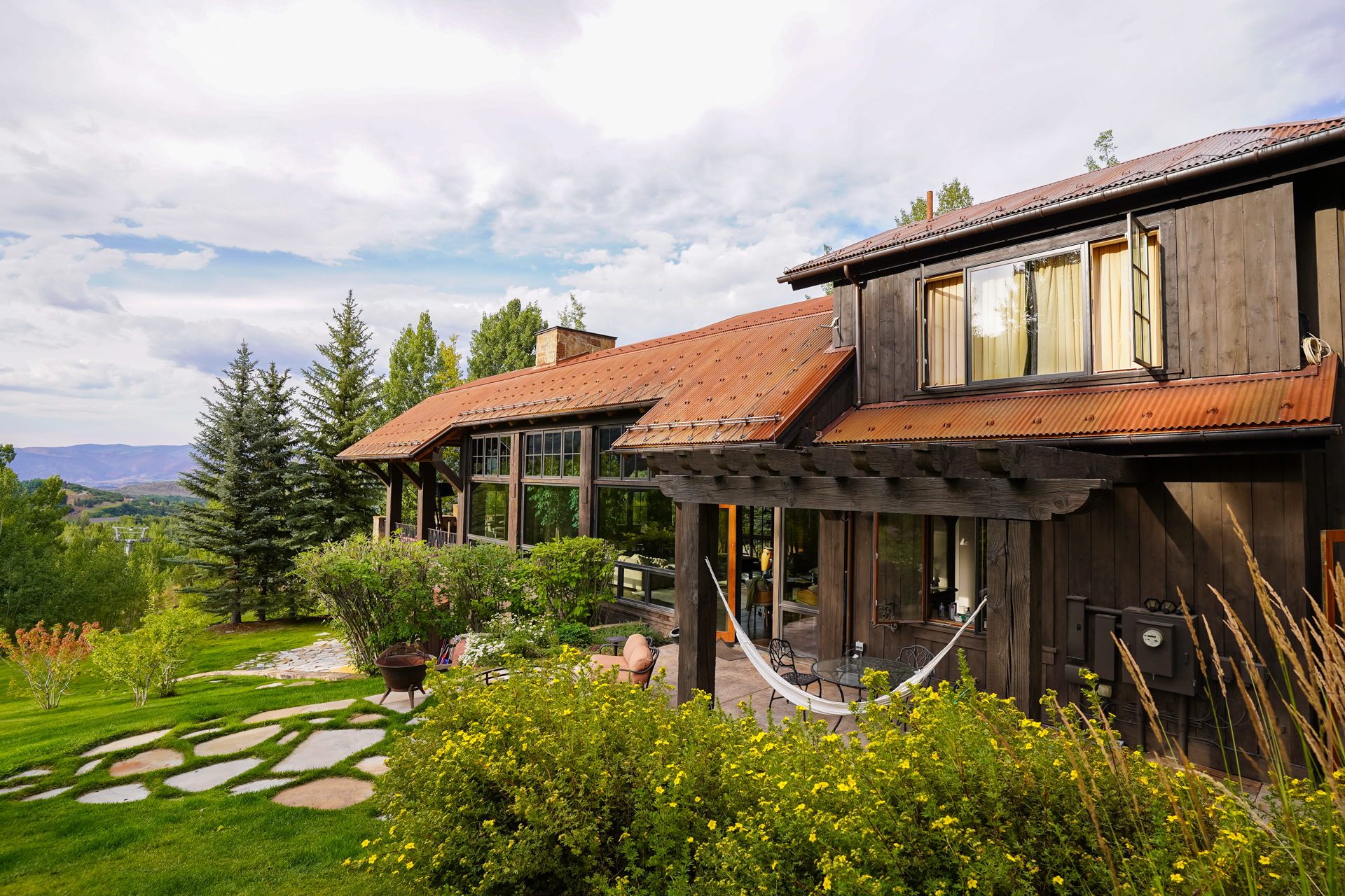 This image showcases the side of a rustic-style home with a weathered metal roof and dark wood siding. A stone pathway leads through a lush green lawn and garden, enhancing the property's curb appeal. A hammock is visible on the patio, suggesting a relaxing outdoor living space.