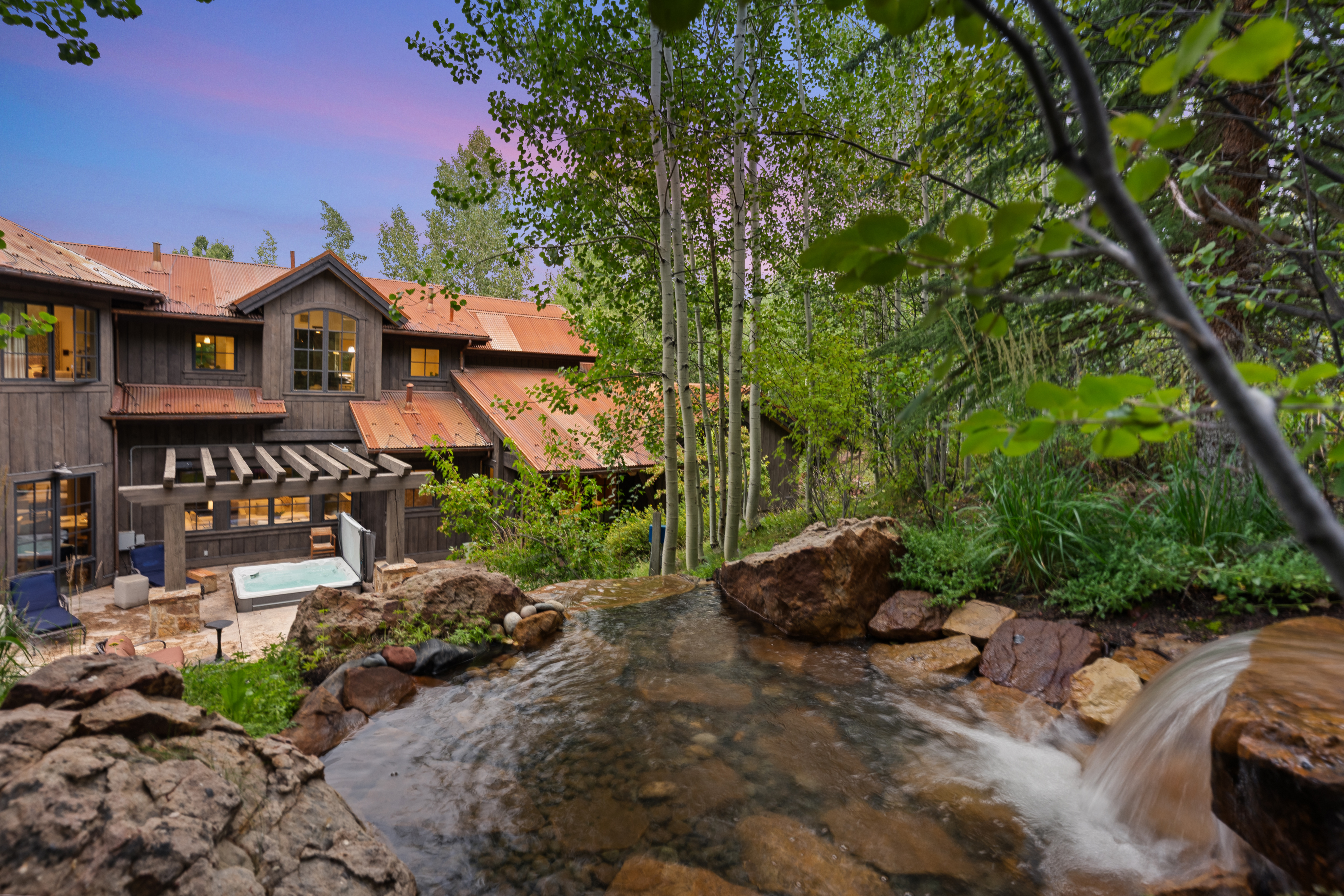 This image showcases the rear exterior of a luxurious mountain home, featuring a rustic design with dark wood siding and a copper roof. A stone waterfall feature flows in the foreground, leading the eye towards a patio area with a hot tub. The surrounding landscape includes lush greenery and aspen trees, creating a serene and private setting.