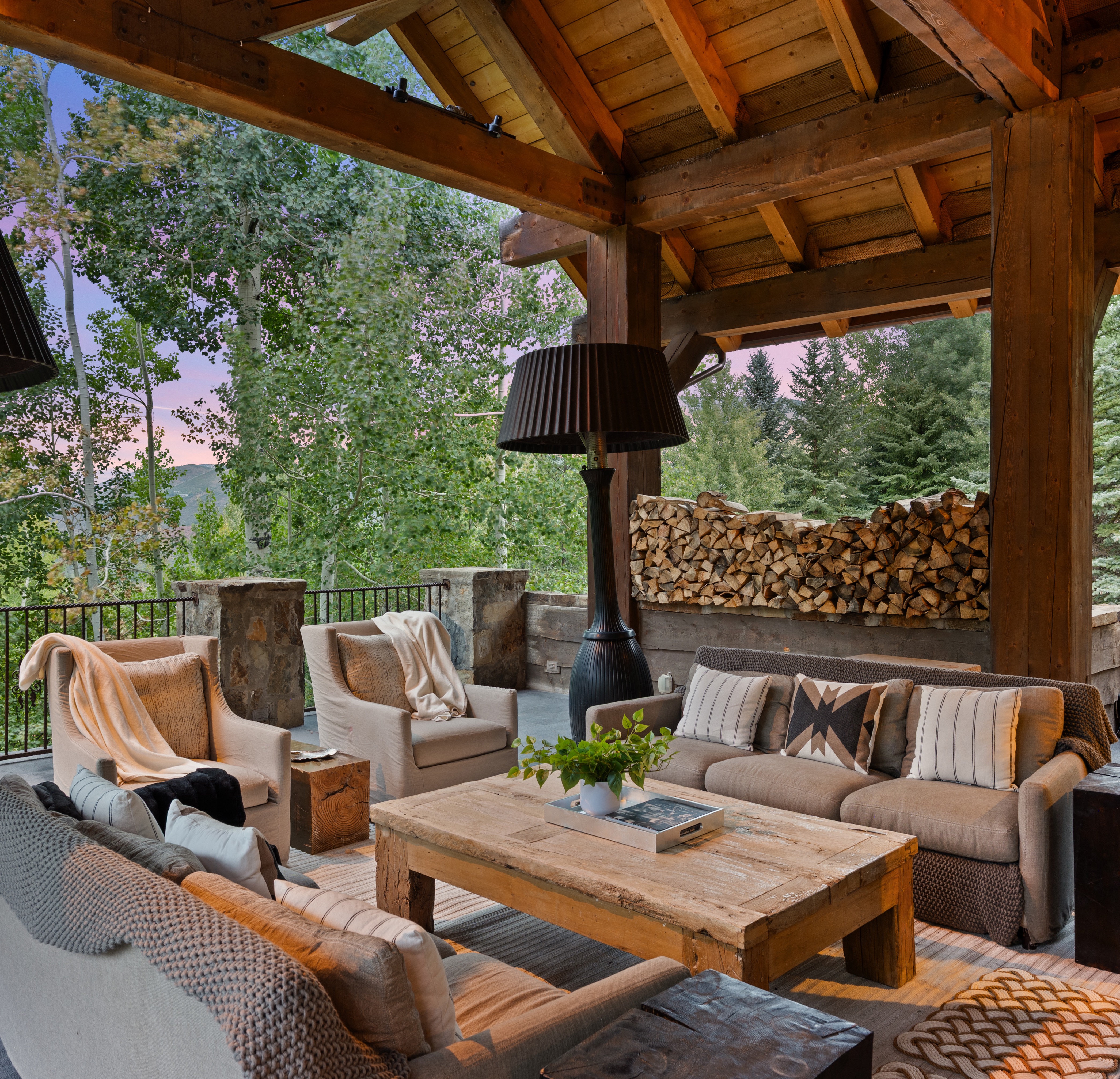 This image showcases a luxurious outdoor living space, featuring comfortable seating arrangements with plush sofas and armchairs, centered around a rustic wooden coffee table. The patio is covered by a wooden structure with exposed beams, creating a cozy and inviting atmosphere. A stone fireplace and stacked firewood add to the rustic charm, while the surrounding trees provide privacy and a scenic backdrop.