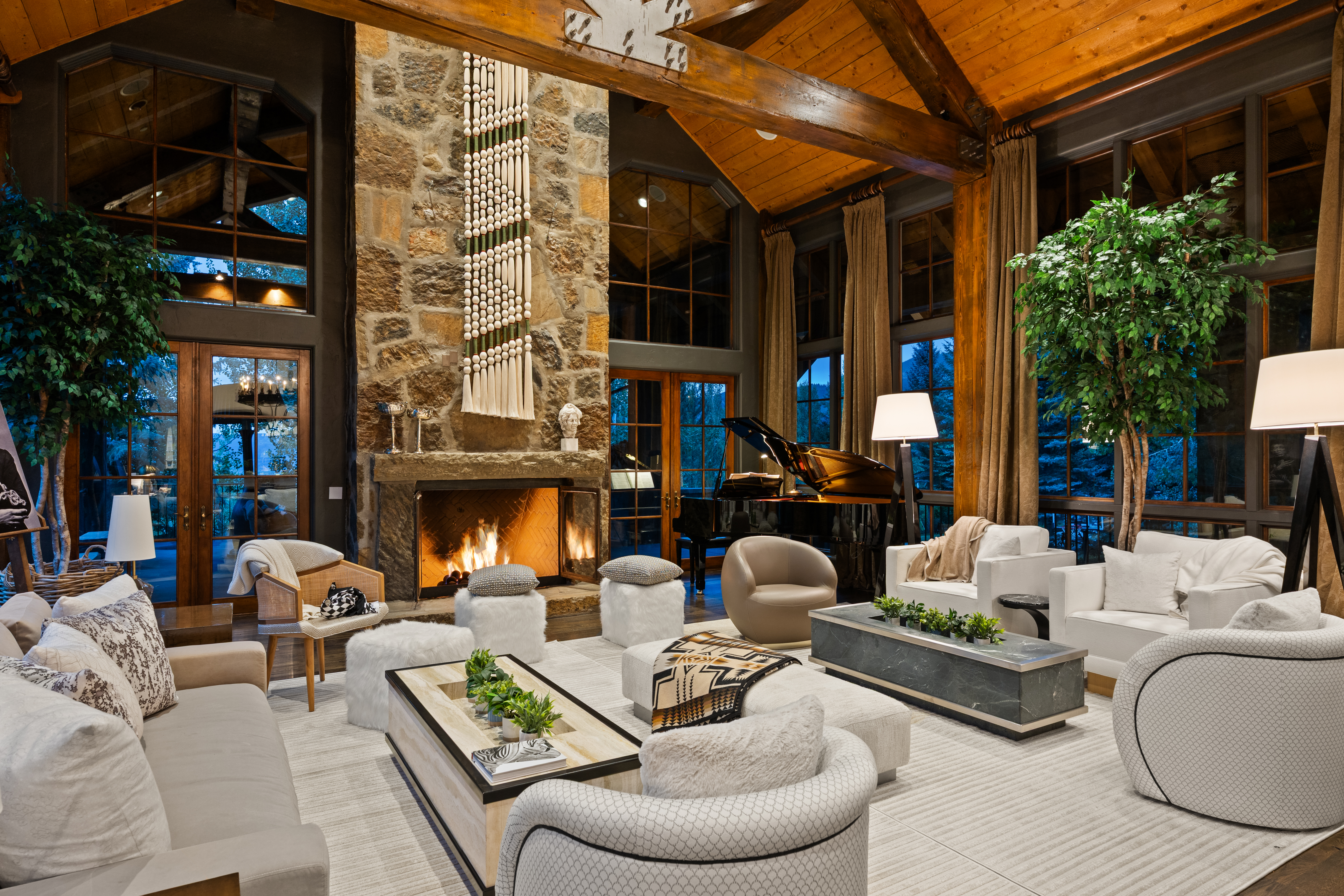 This is a luxurious living room featuring a stone fireplace with a decorative hanging tapestry, a grand piano, and comfortable seating arrangements. The room has high ceilings with exposed wooden beams and large windows that offer natural light. The overall style is elegant and inviting, perfect for entertaining or relaxing.
