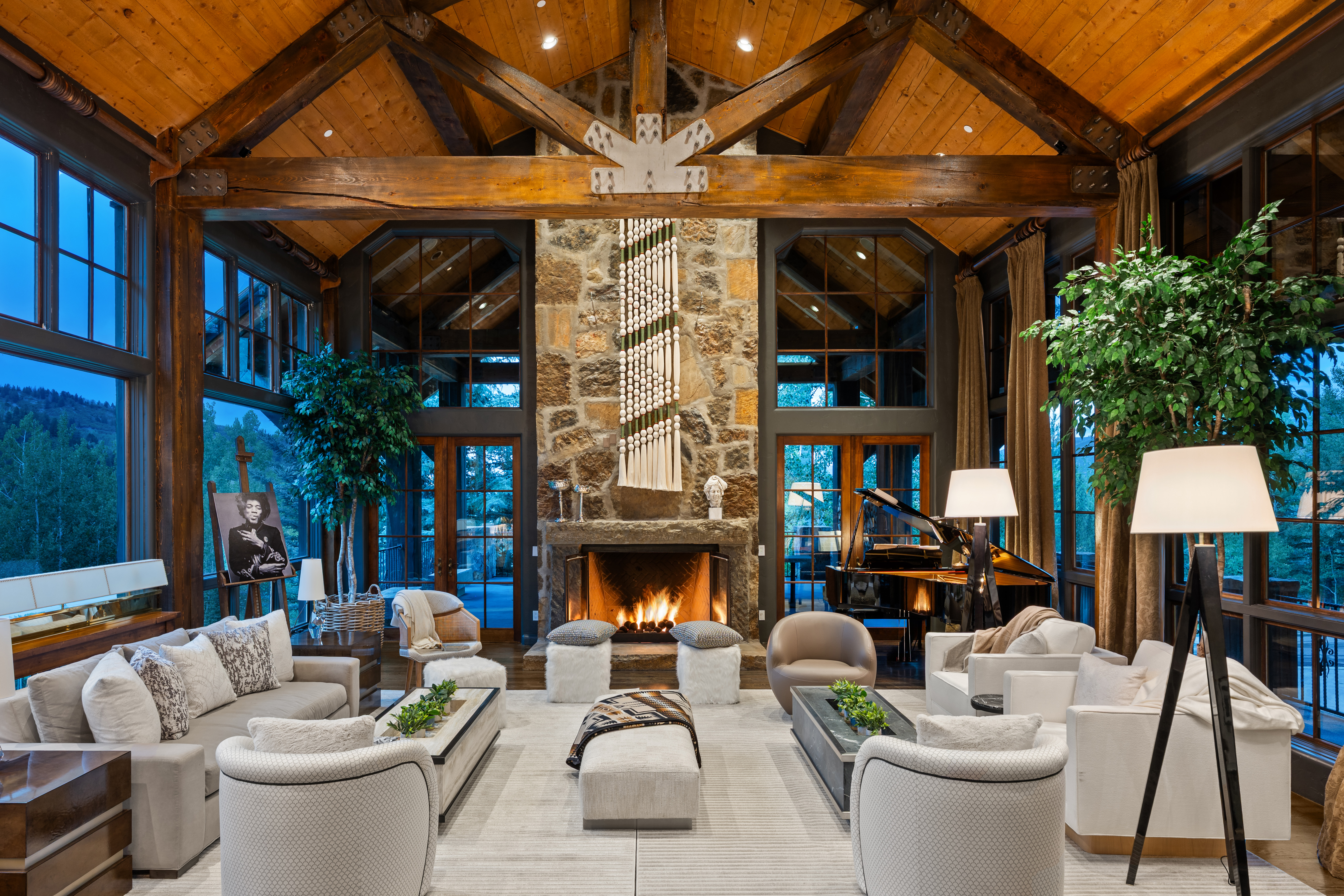 This is a luxurious living room featuring a high vaulted ceiling with exposed wooden beams and a grand stone fireplace. The room is furnished with plush sofas, armchairs, and a grand piano, creating an inviting and sophisticated atmosphere. Large windows offer views of the surrounding landscape, enhancing the room's spacious and elegant feel.
