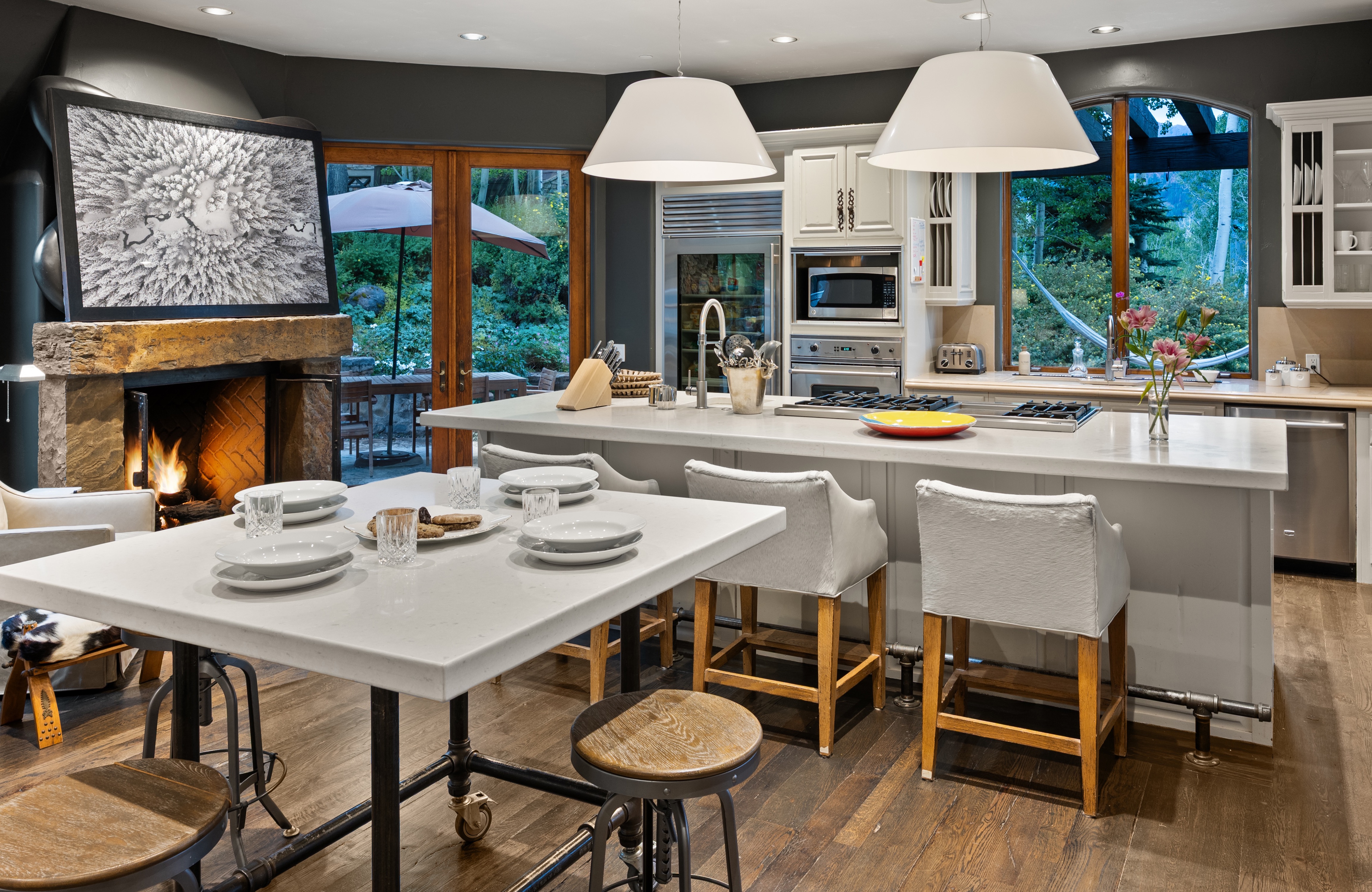 This is a well-lit, inviting kitchen featuring a large island with seating, white cabinetry, and stainless steel appliances. A stone fireplace with a large framed picture adds a unique, cozy touch to the space. The open layout and view to the outside patio create a seamless indoor-outdoor living experience.
