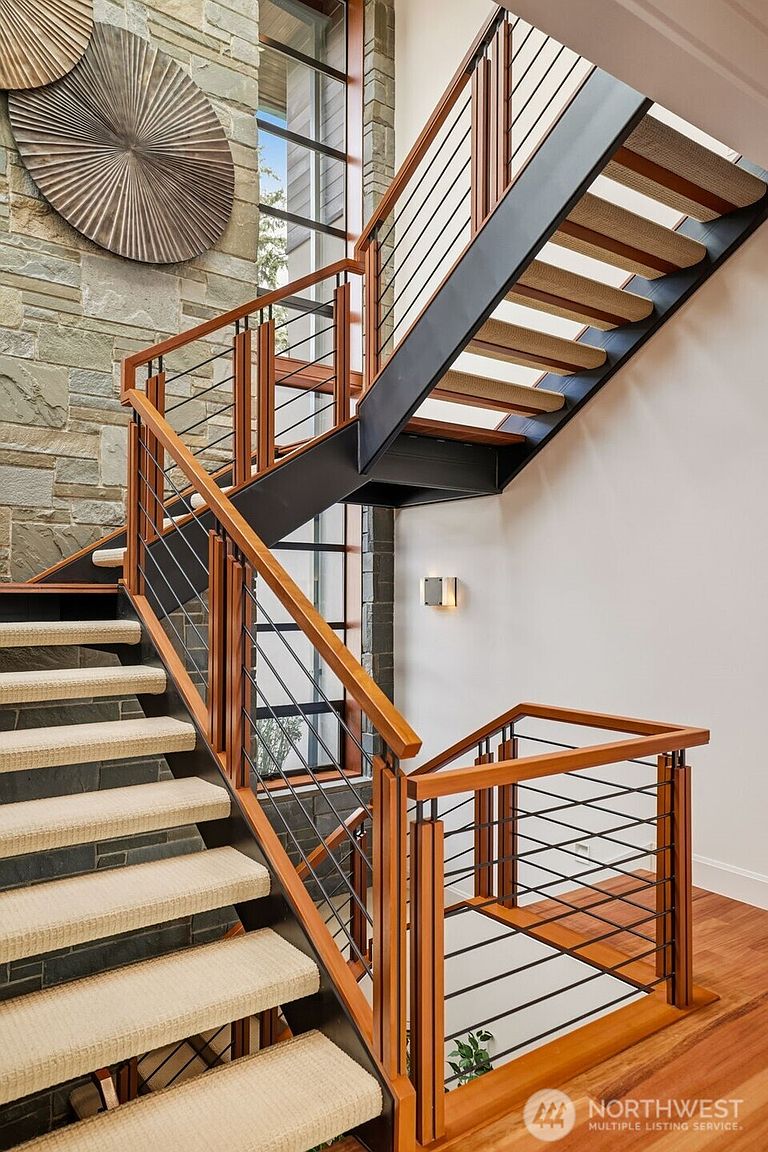 This striking staircase features a modern design with a black metal frame, warm wood handrails, and horizontal cable railings. The steps are finished with light-colored carpet runners, contrasting against the stone-clad wall that rises alongside the stairs. A large window assembly provides natural light, while decorative circular wall art adds a sophisticated touch to the landing area.