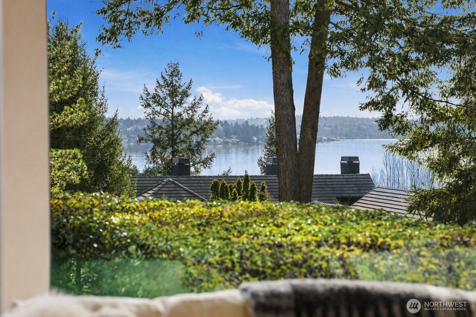 This view from inside a home captures a serene, elevated perspective looking out over a lush green hedge toward a calm body of water. In the foreground, the blurred texture of a soft furnishing suggests a cozy interior setting, while the mid-ground features the dark shingled roofs of neighboring homes nestled among mature trees. The scene is framed by the natural beauty of the outdoors, emphasizing a peaceful, scenic atmosphere perfect for relaxation.